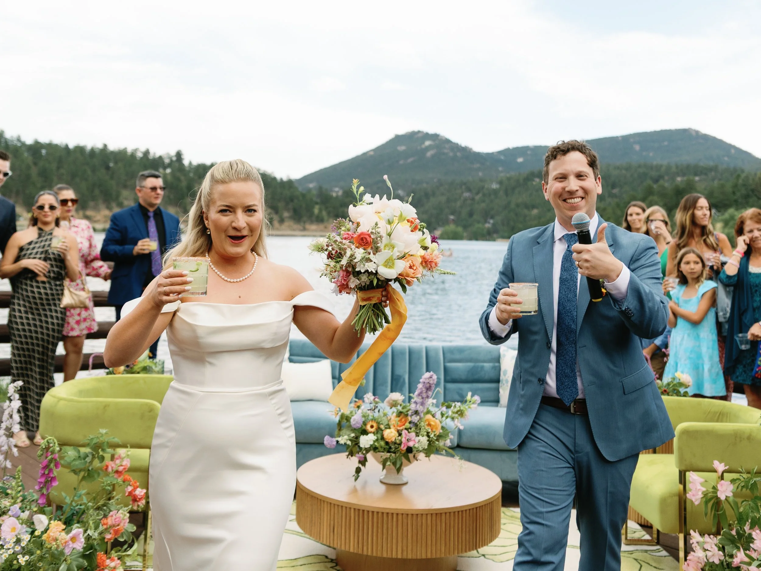 Bride and groom holding drinks and celebrating outdoors by a lake with mountains in the background. The bride holds a bouquet of flowers, and the groom has a microphone. Guests stand behind them, some in colorful dresses and suits, smiling and watchi