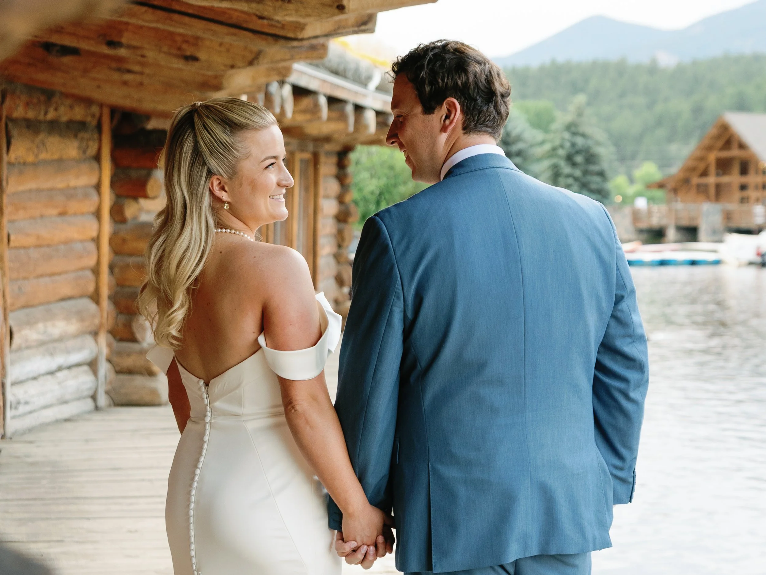 A bride and groom hold hands and look at each other, standing outside near a lake with mountains in the background, in front of a wooden cabin.