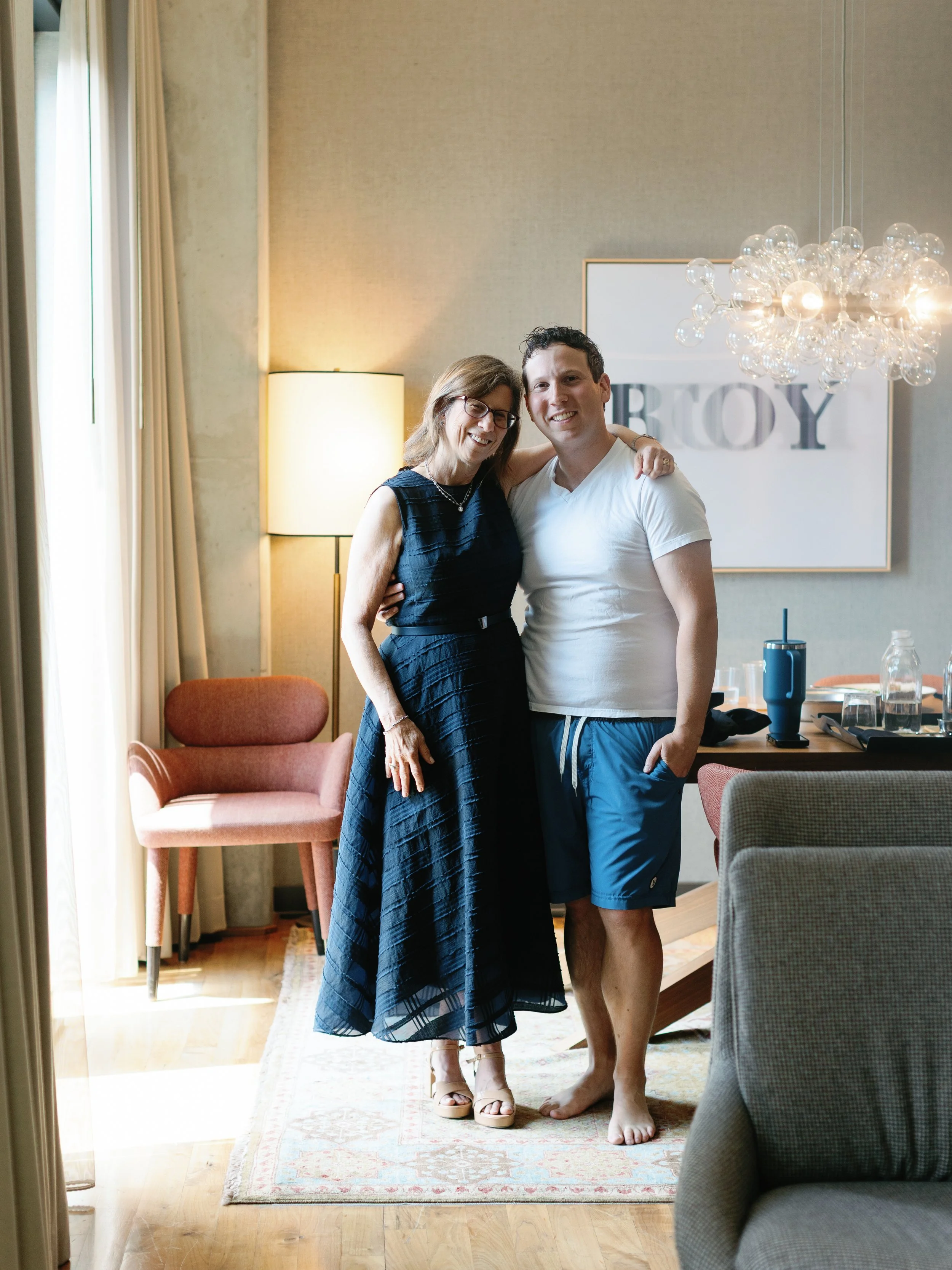 A woman and a young man embrace and smile at the camera inside a living room with natural light. The woman wears a black dress and glasses, while the young man wears a white T-shirt and blue shorts. Behind them is a wall with a framed poster that say