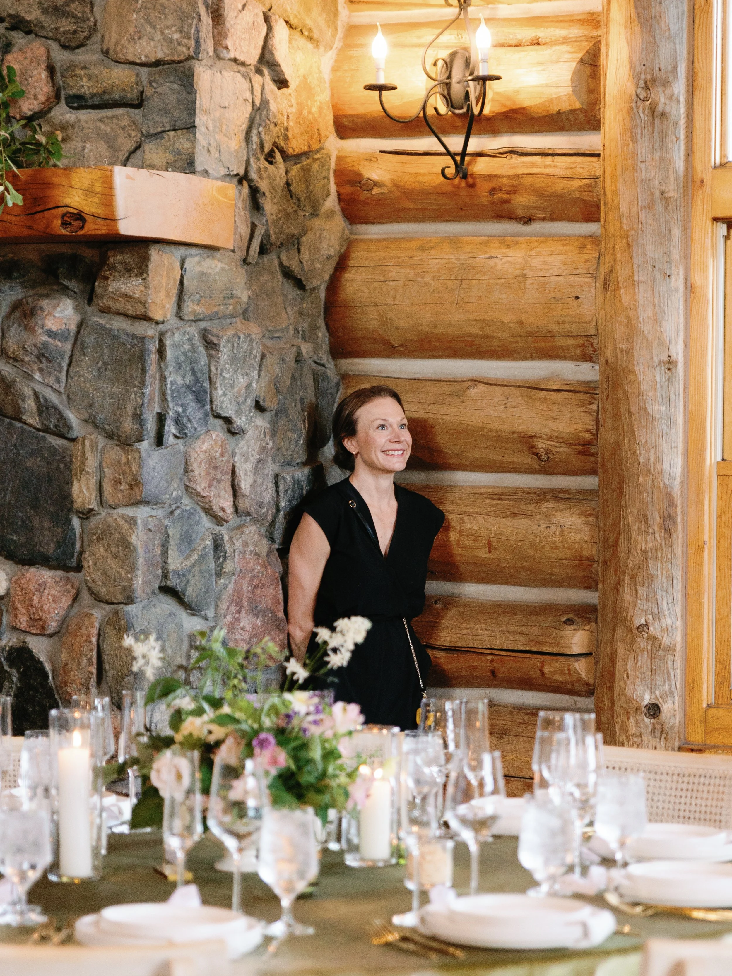 A woman smiling and standing behind a decorated dining table in a rustic wooden room with a stone wall and a chandelier.