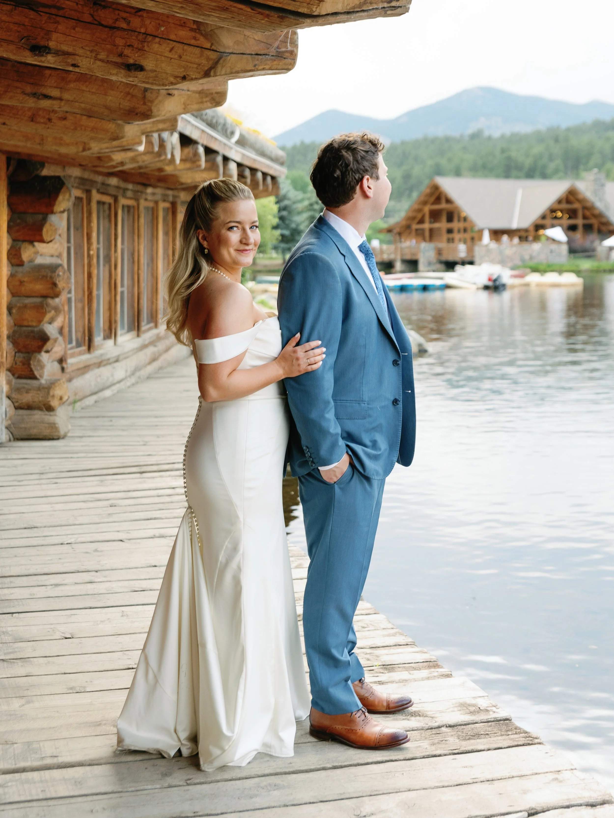 A couple dressed in formal wedding attire standing on a wooden dock by a lake, with mountains and log cabins in the background, during daytime.