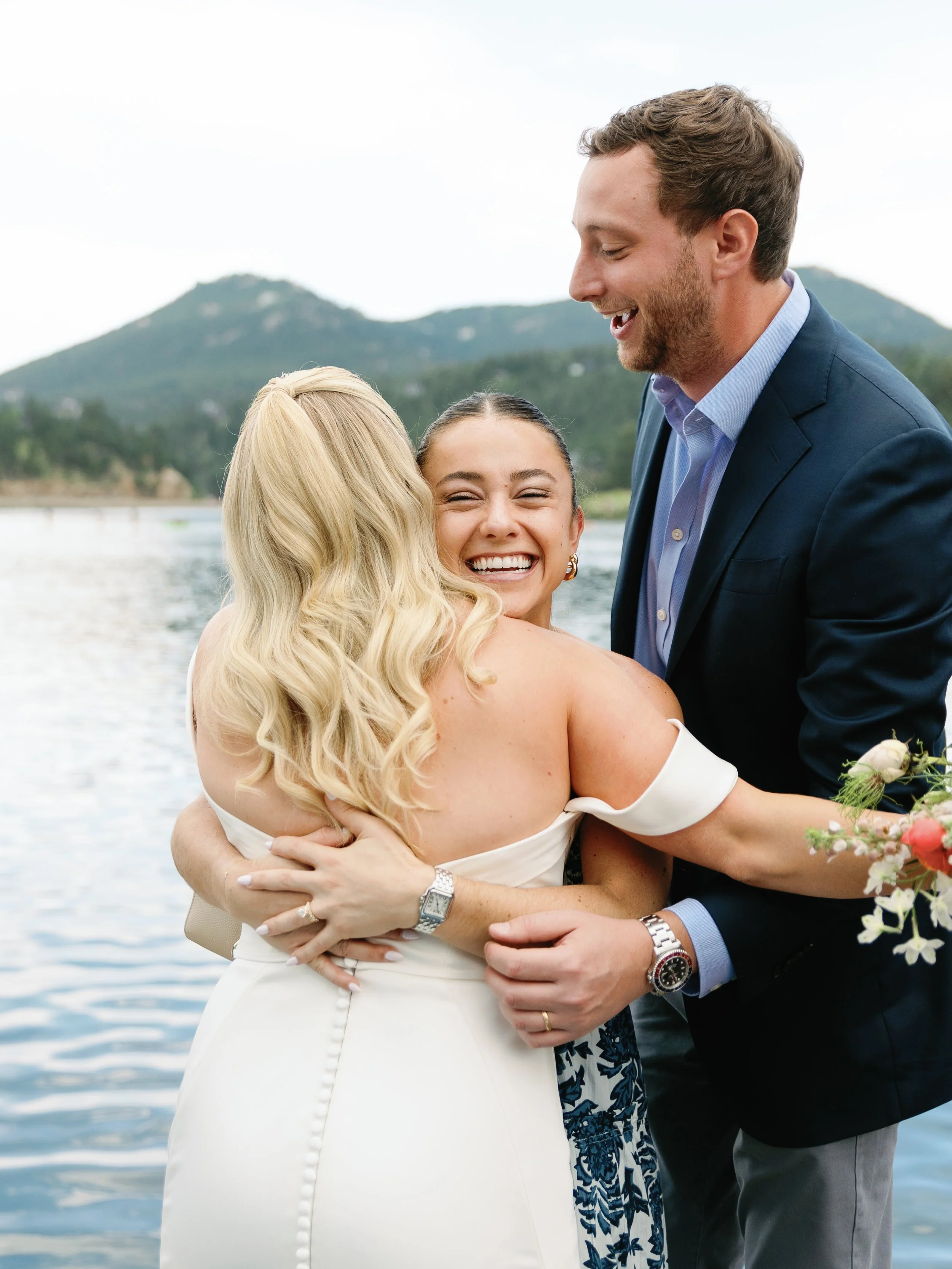 A happy group of three people hugging outdoors near a body of water with mountains in the background; the woman in the center is smiling broadly, with two other individuals, one in a white dress and the other in a dark suit, embracing her.