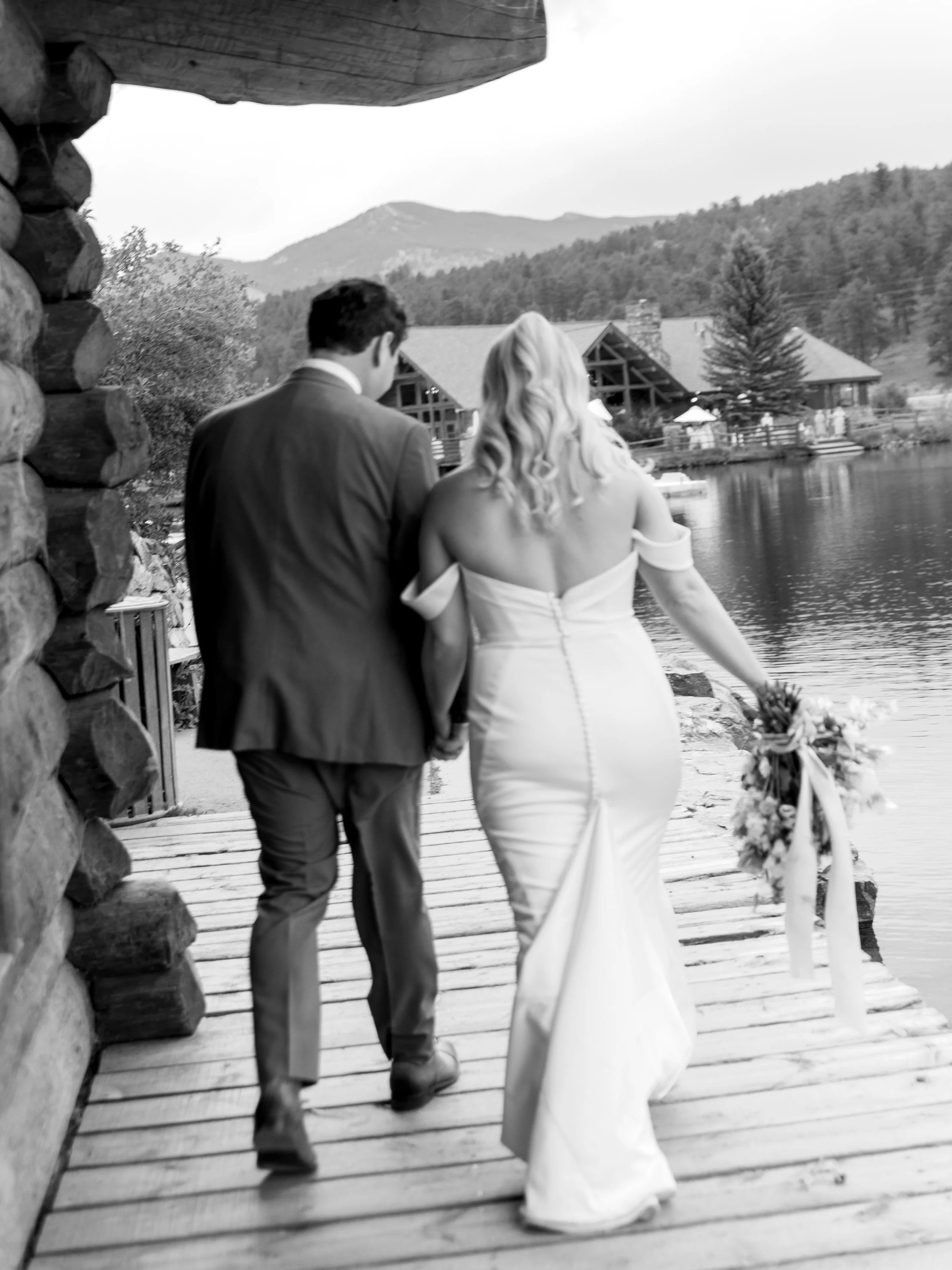 A black and white photo of a bride and groom walking hand in hand on a wooden dock beside a lake, with mountains and a cabin in the background.