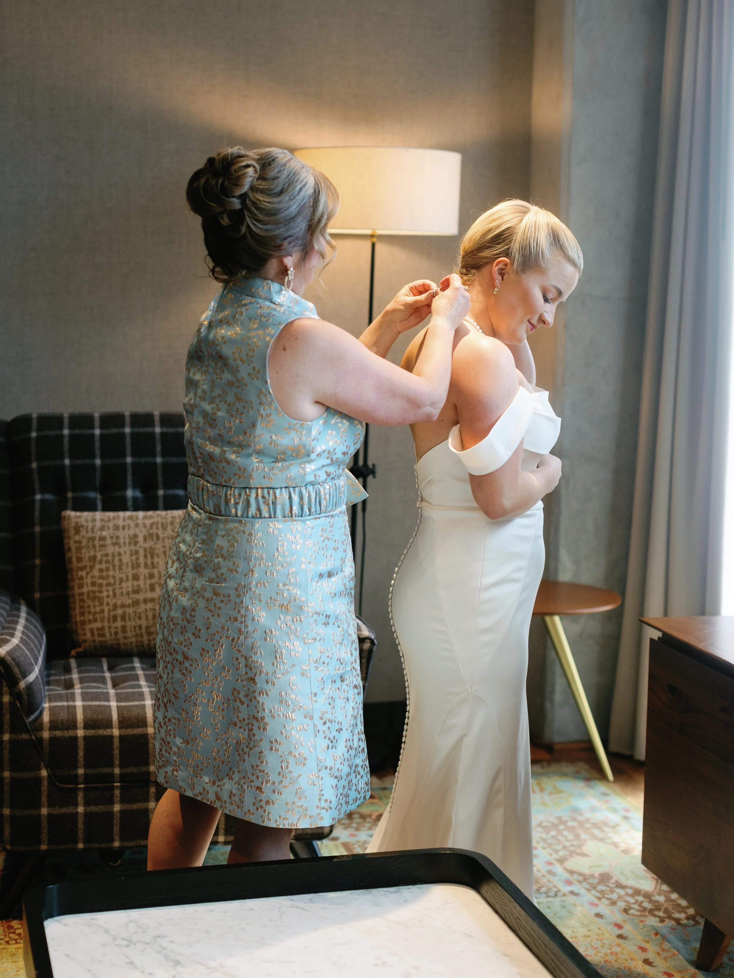 A woman helps another woman put on earrings in a hotel room, with a lamp and curtains in the background.