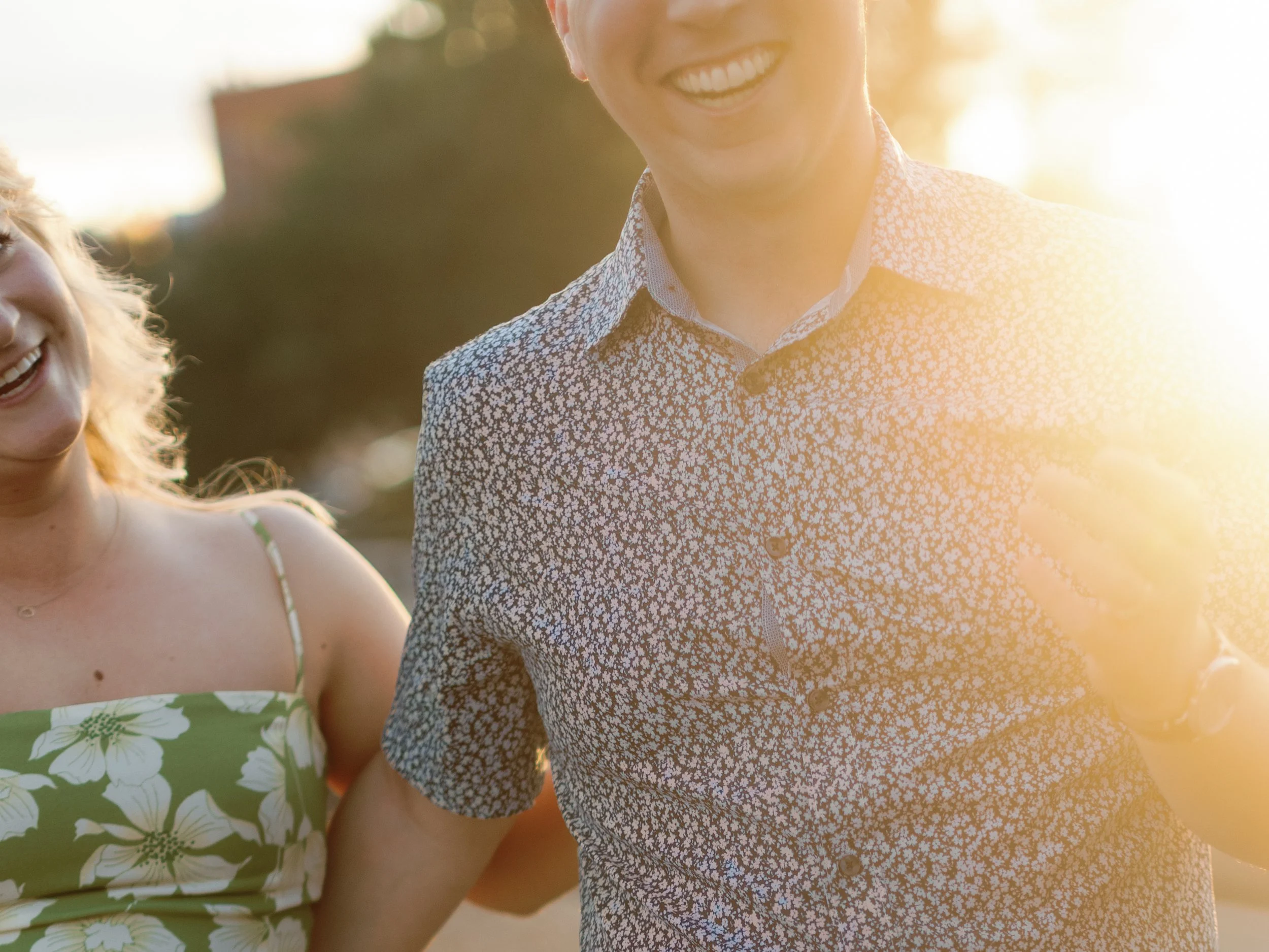 A smiling woman in a green floral dress and a smiling man in a patterned shirt are outside during sunset, enjoying each other's company.