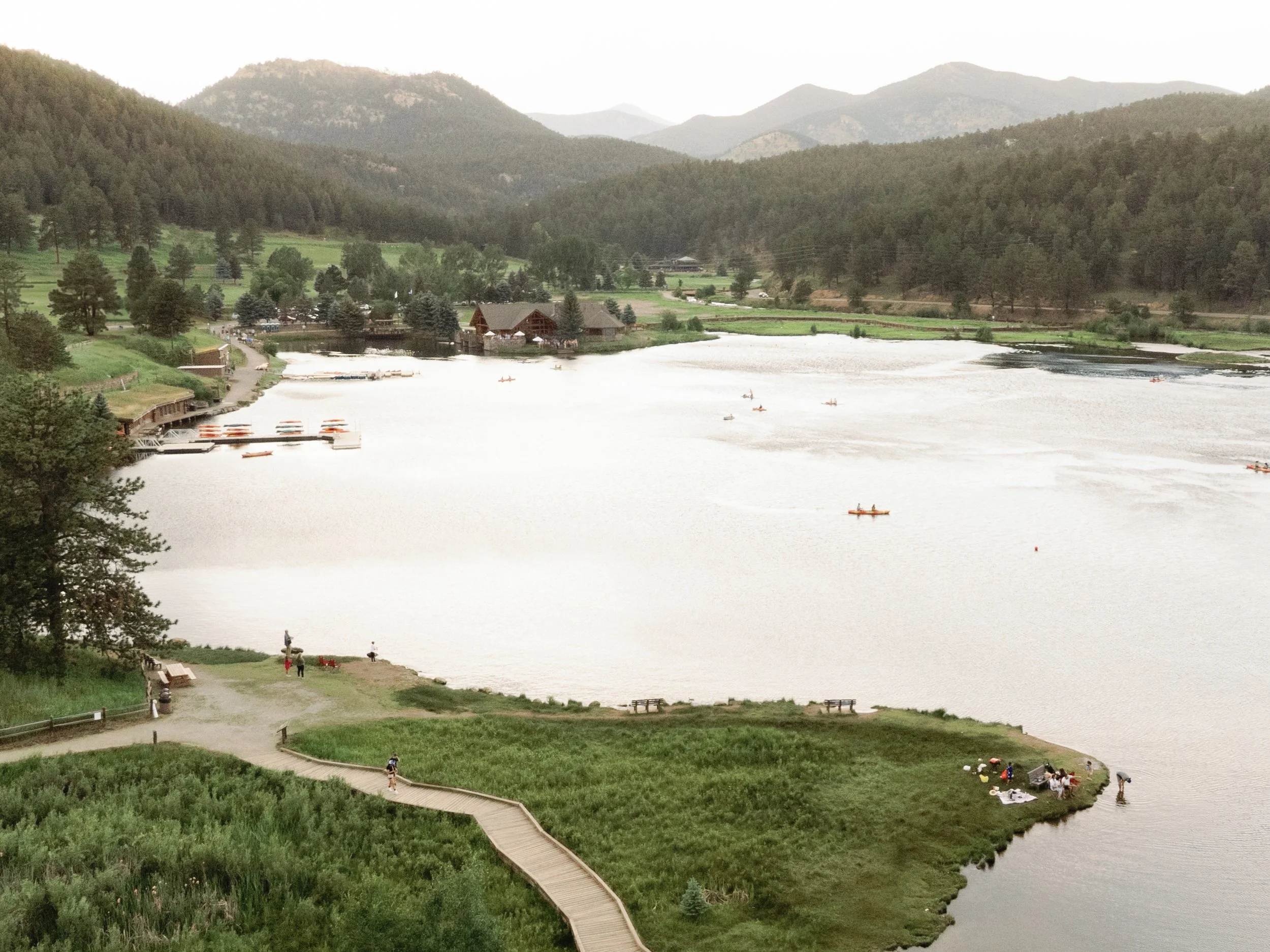 View of a calm lake surrounded by green hills and mountains with some boats on the water, a wooden path leading to a small grassy area with people, trees, and houses in the background.