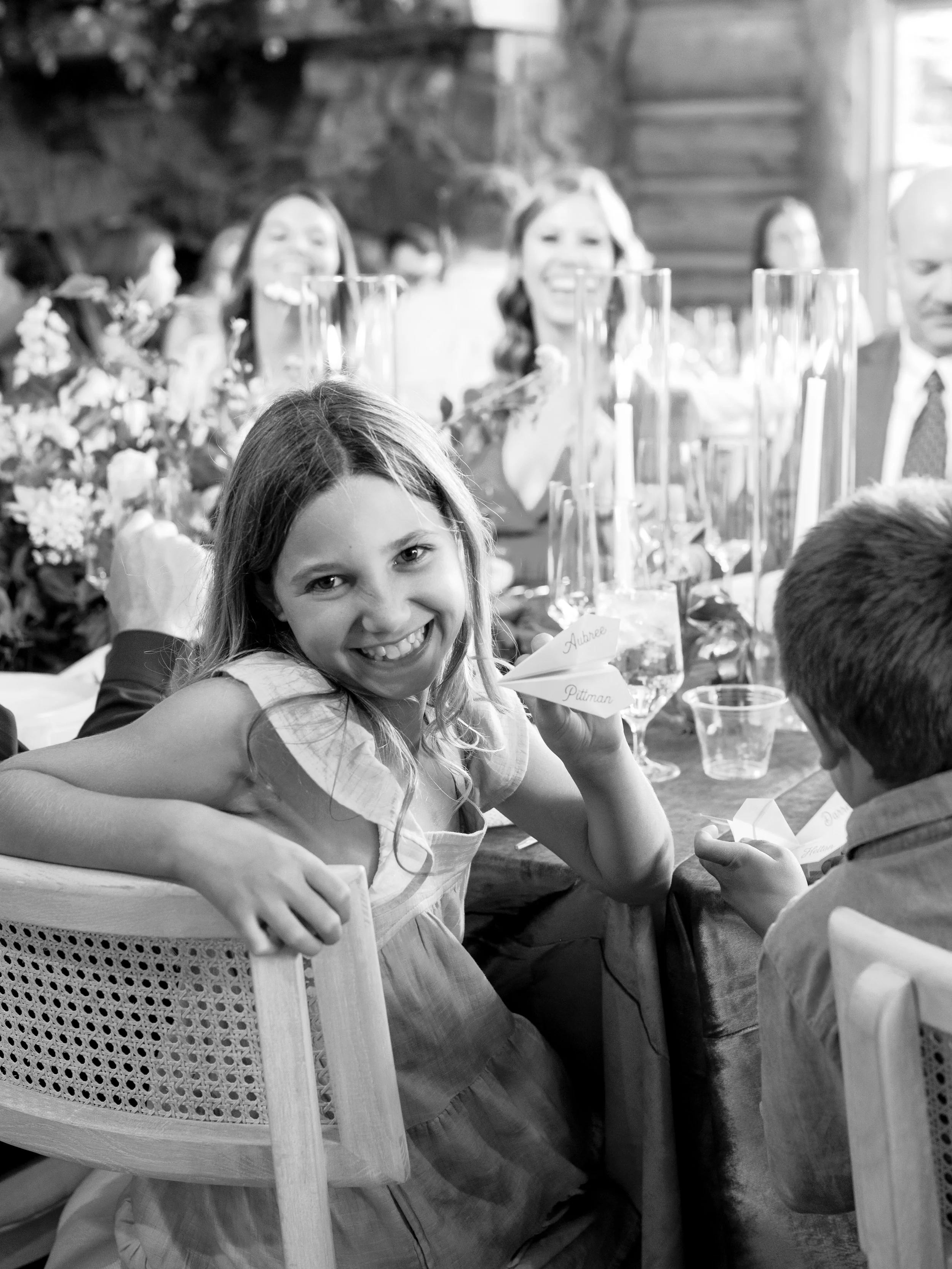 A young girl smiling and holding a paper airplane with the name 'Aubrey Putman' written on it, sitting at a decorated table during a celebration or event, with other people and floral arrangements in the background.