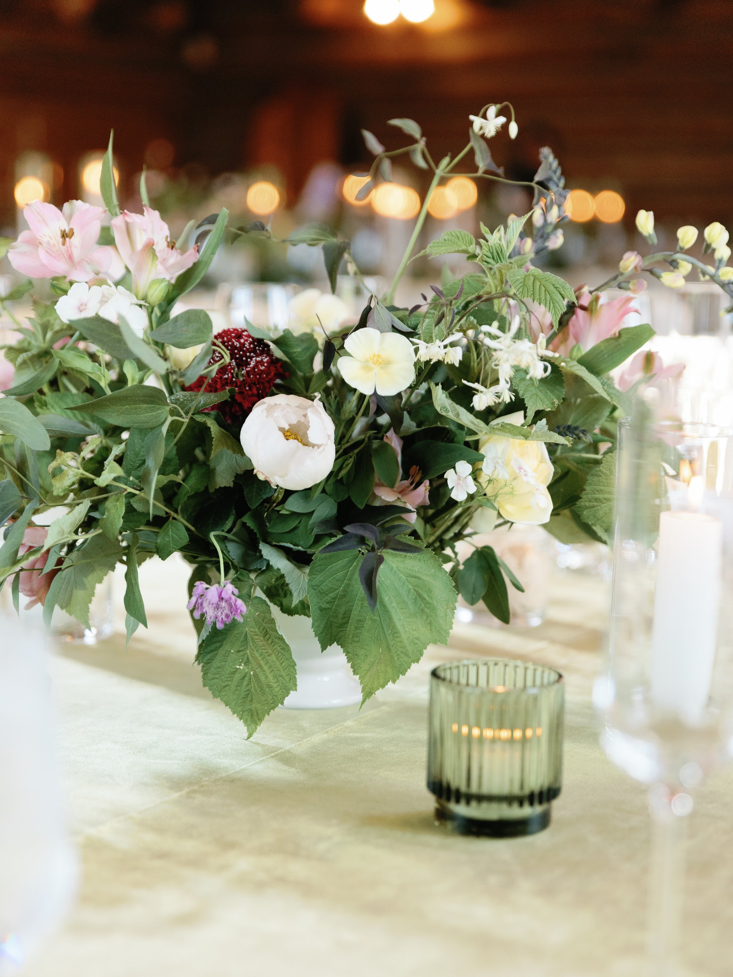 A bouquet of mixed flowers including pink lilies, white roses, and purple flowers arranged on a table with a green candle holder and lit candles in the background.