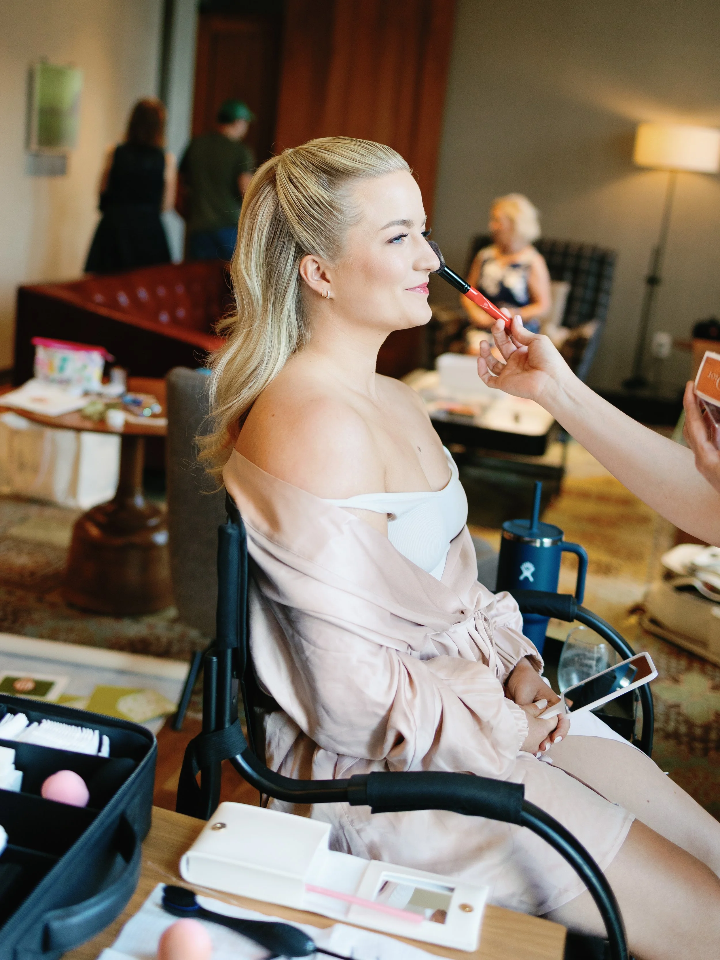 Woman sitting in a chair getting her makeup done, with makeup tools on the table in front of her.