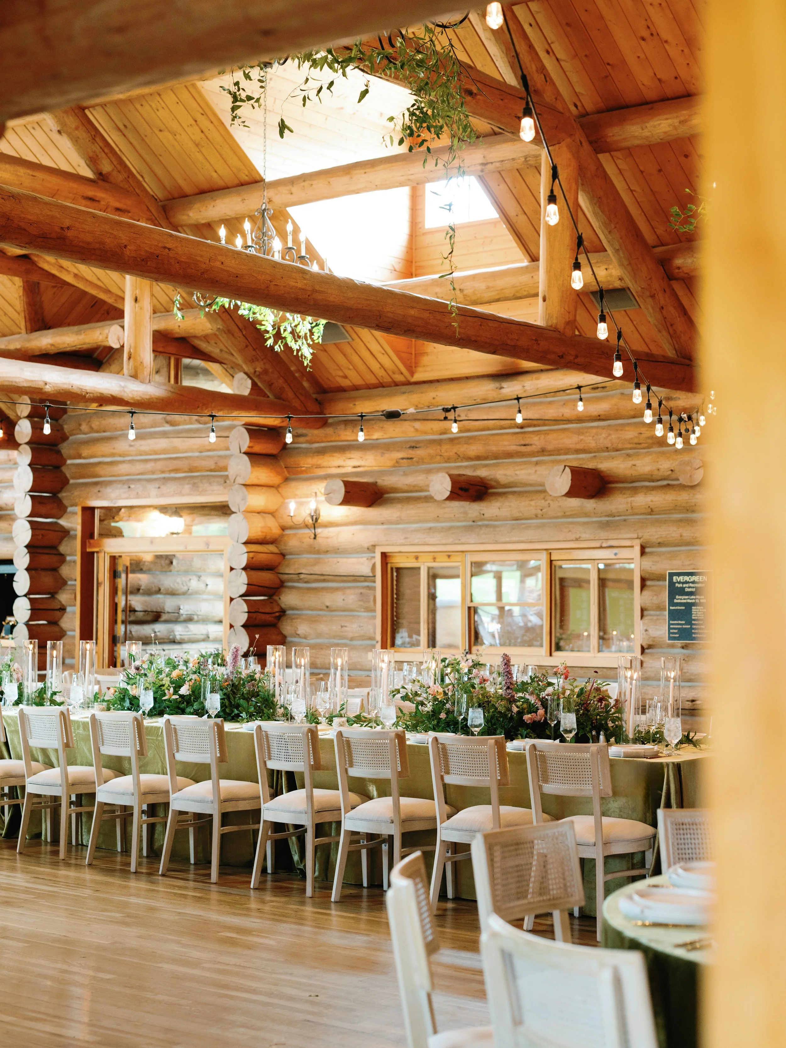 Decorated banquet table in a rustic log cabin with string lights and floral centerpieces, overlooking a wood-paneled ceiling and windows.