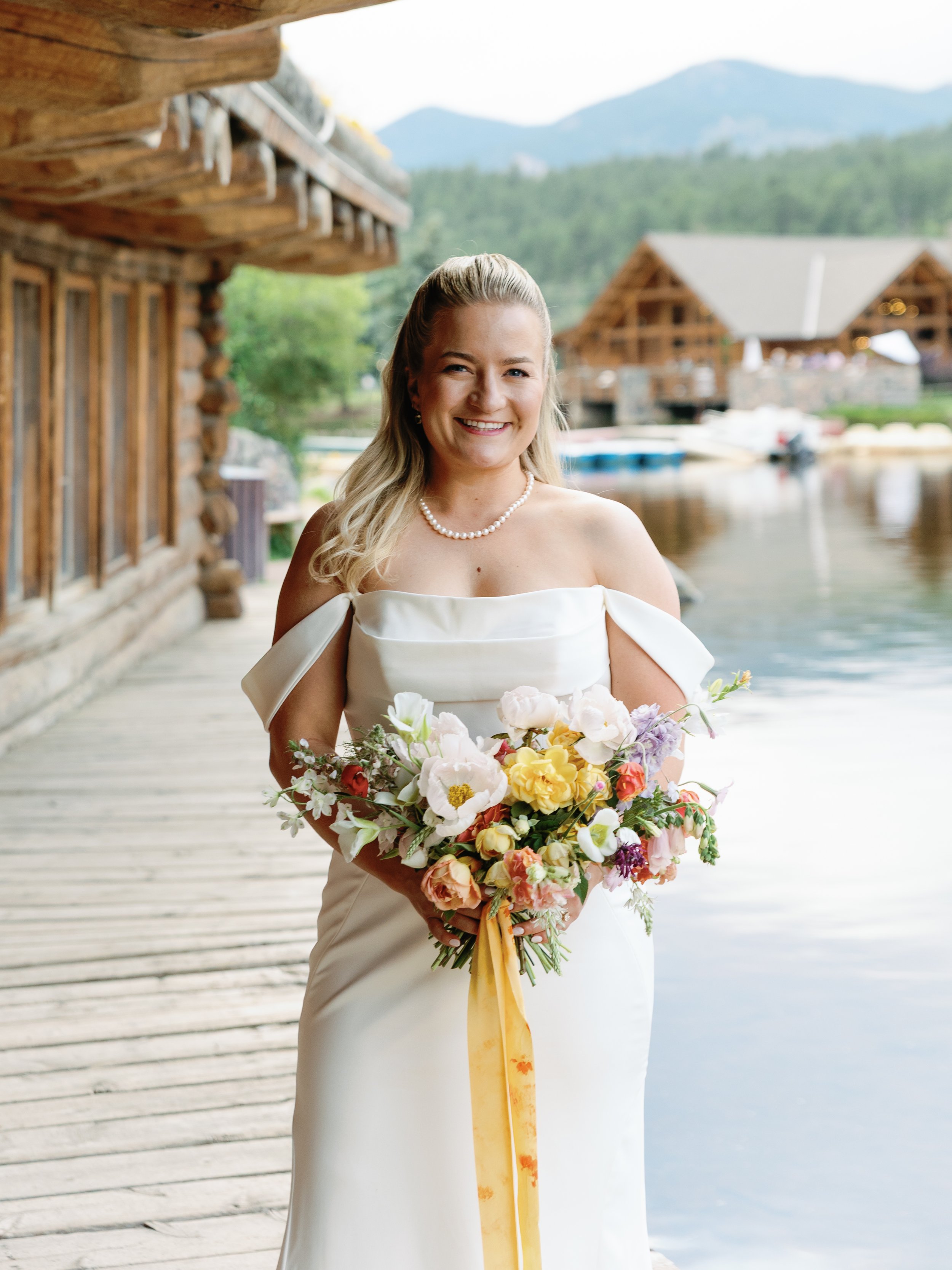 A smiling bride in a white wedding dress holding a colorful bouquet of flowers standing on a wooden dock by a lake with mountains and wooden cabin-like structures in the background.