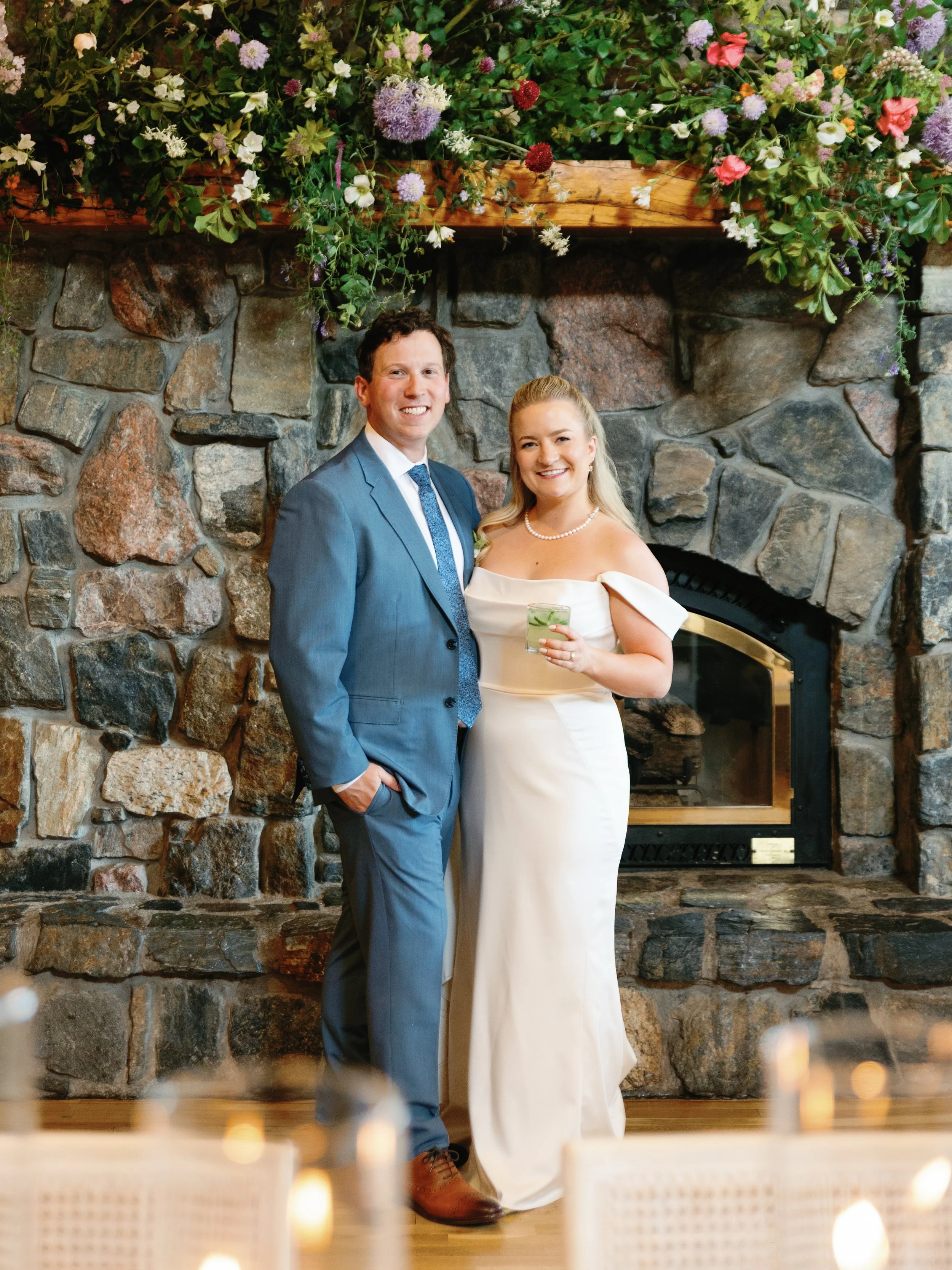 A couple in wedding attire standing in front of a stone fireplace decorated with flowers, smiling at the camera. The groom is wearing a blue suit and tie, while the bride is in a white wedding dress holding a drink.