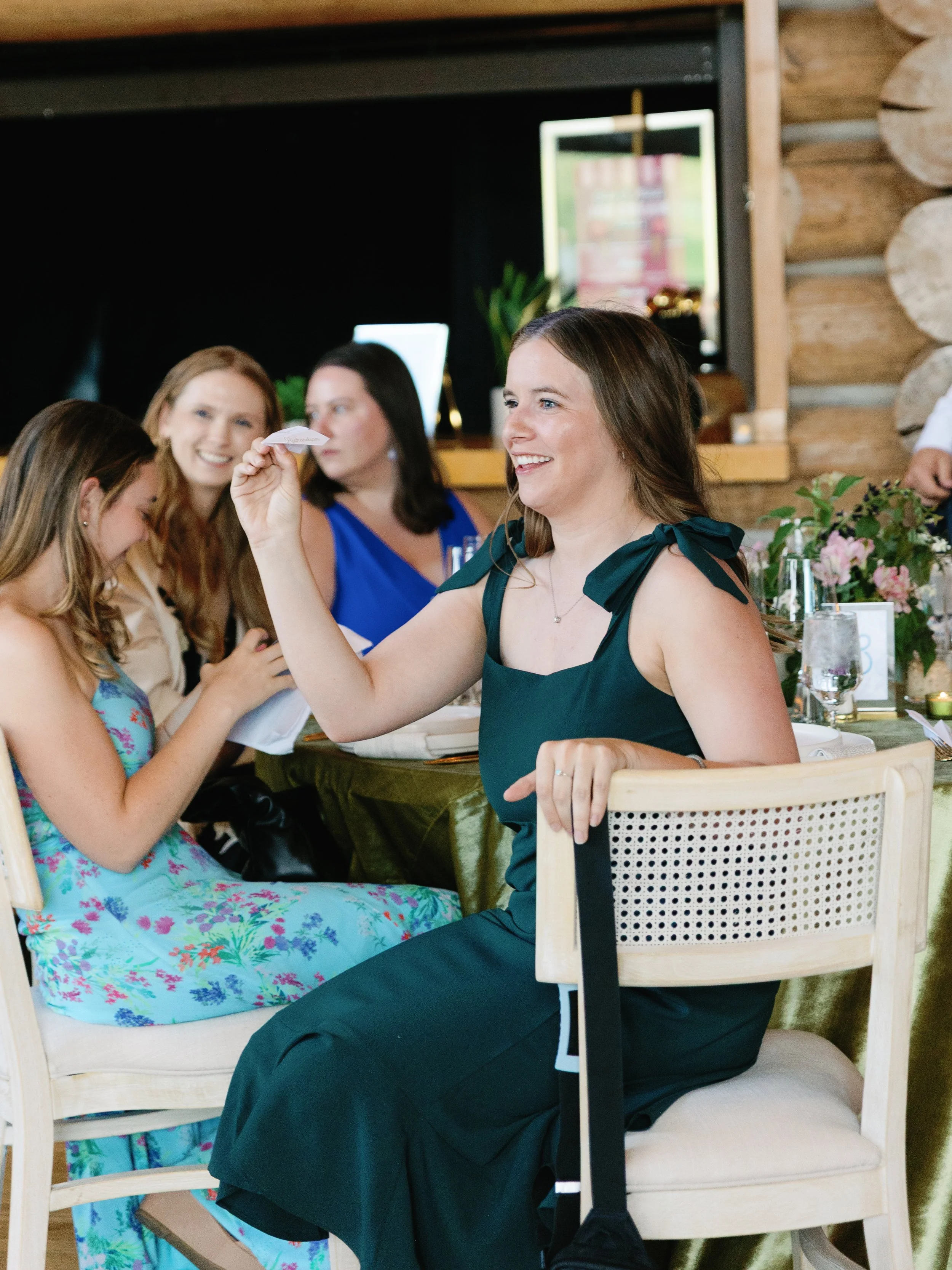 A woman in a dark green dress is sitting at a table, smiling, and holding a small paper item. There are other women around the table, some smiling and looking at her. The setting appears to be a celebration or gathering in a rustic indoor space with 