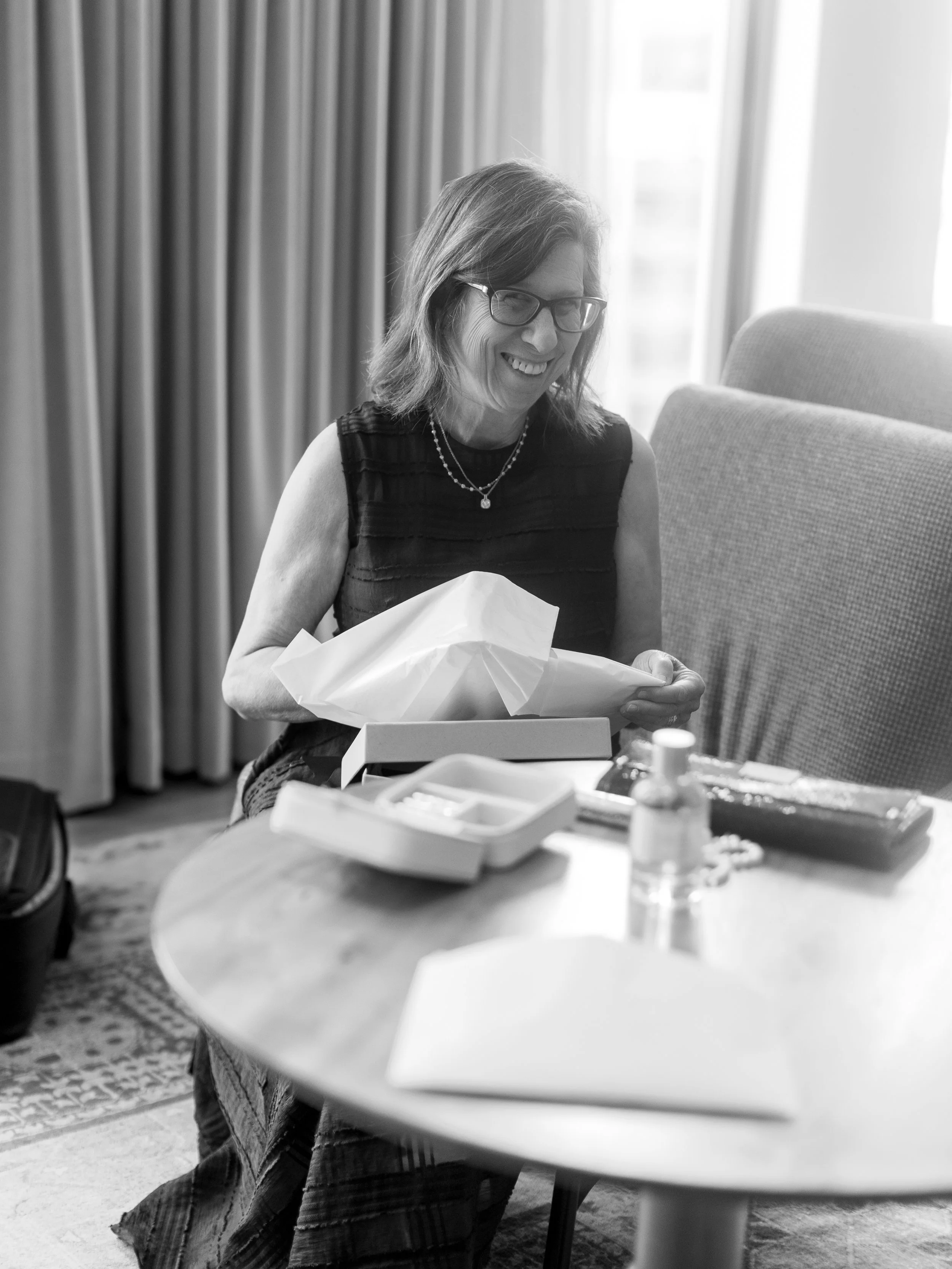 A woman with glasses unwrapping a gift at a table with various items, smiling.