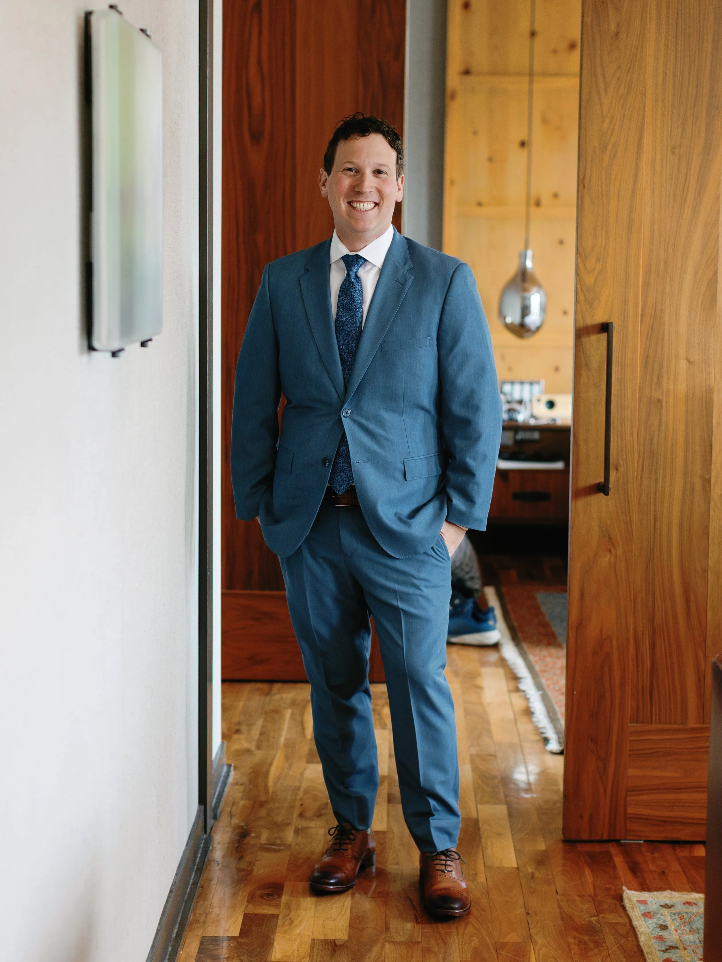 A man in a blue suit and patterned tie, smiling, standing inside a home with wooden floors and walls.