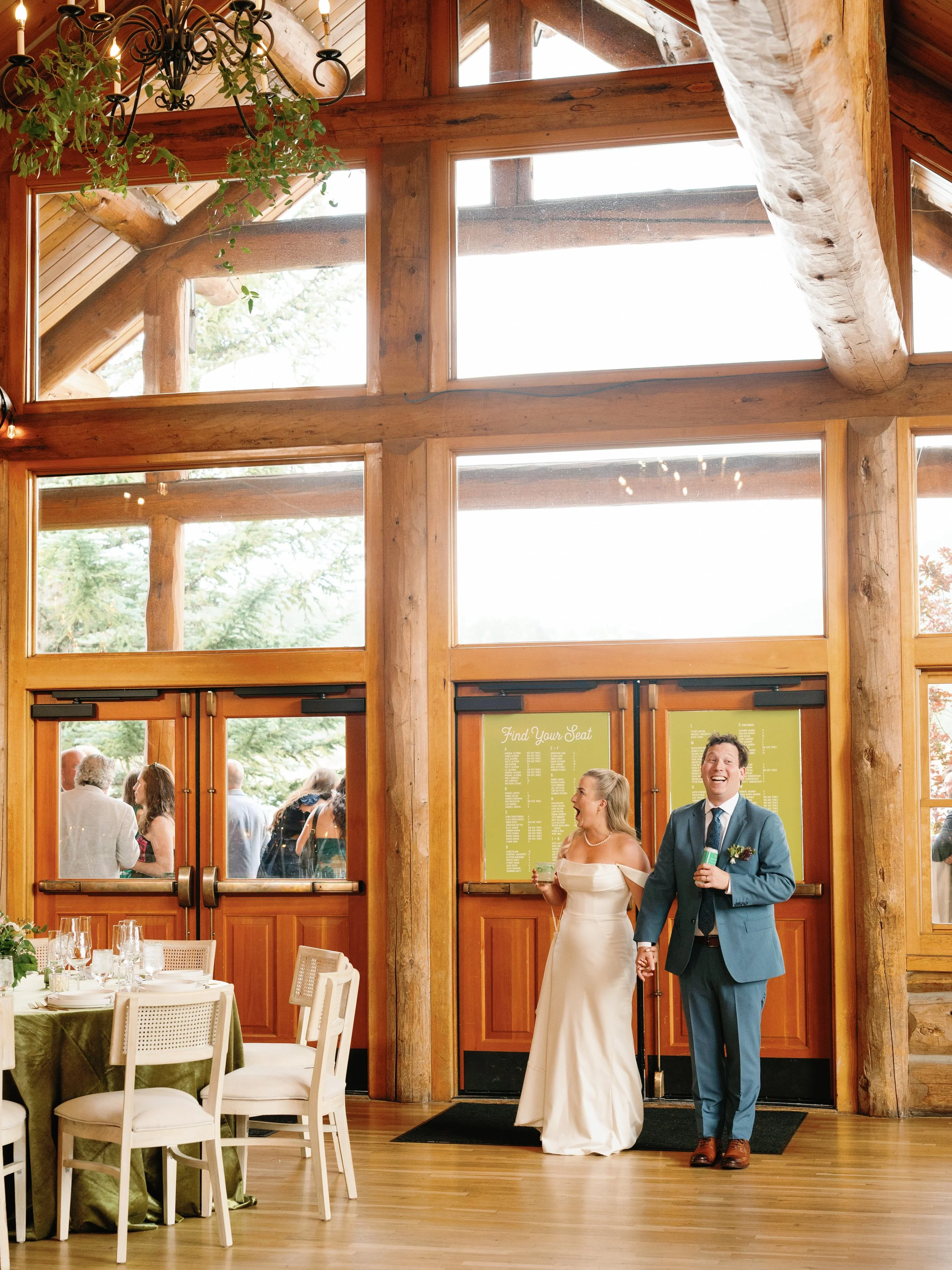 Bride and groom smiling and holding hands inside a rustic wedding venue with wooden beams and large windows, guests visible outside, table with green tablecloth and place settings to the left.