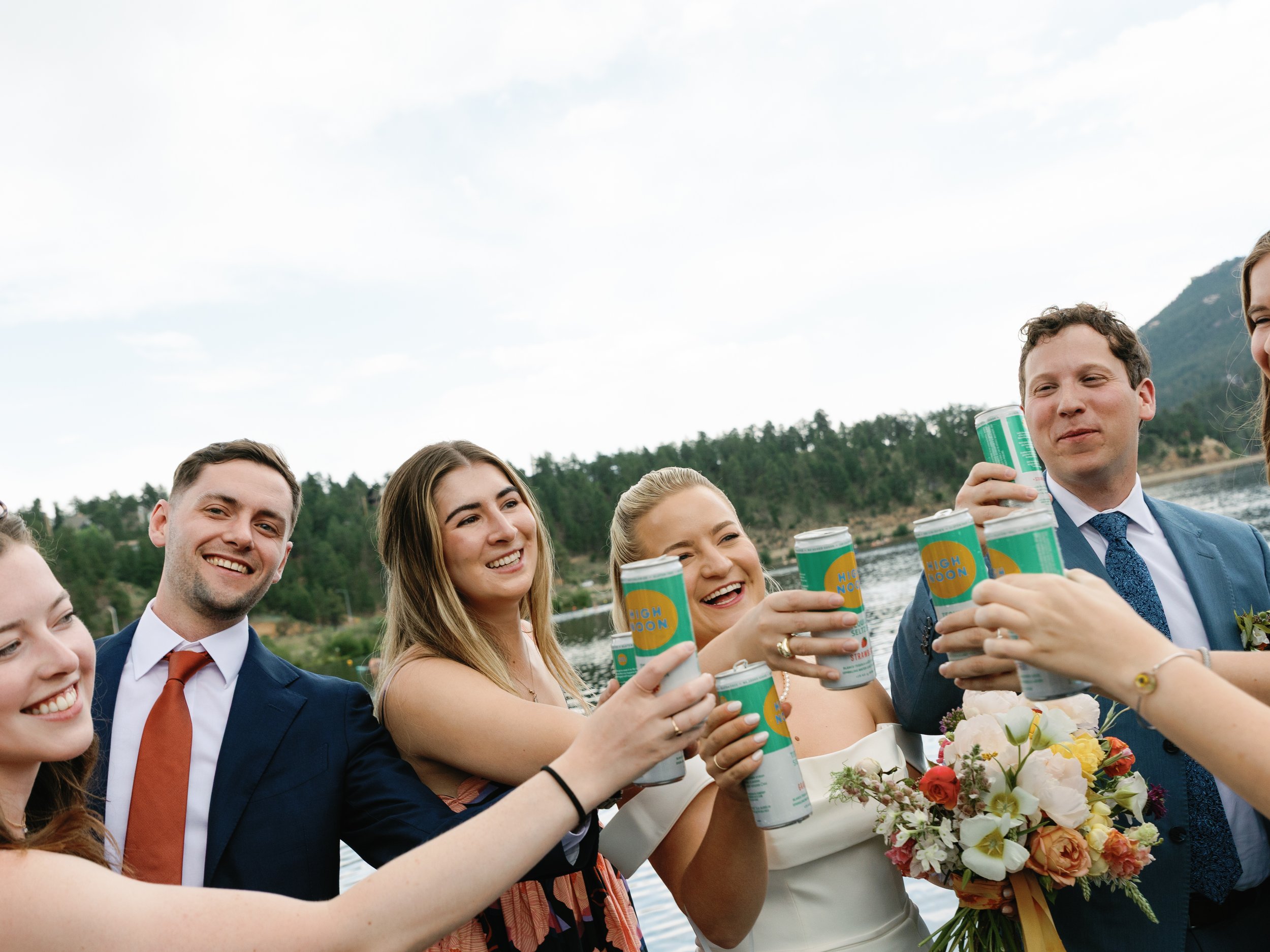 Group of people celebrating outdoors near a lake, raising canned drinks, with trees and mountains in the background.