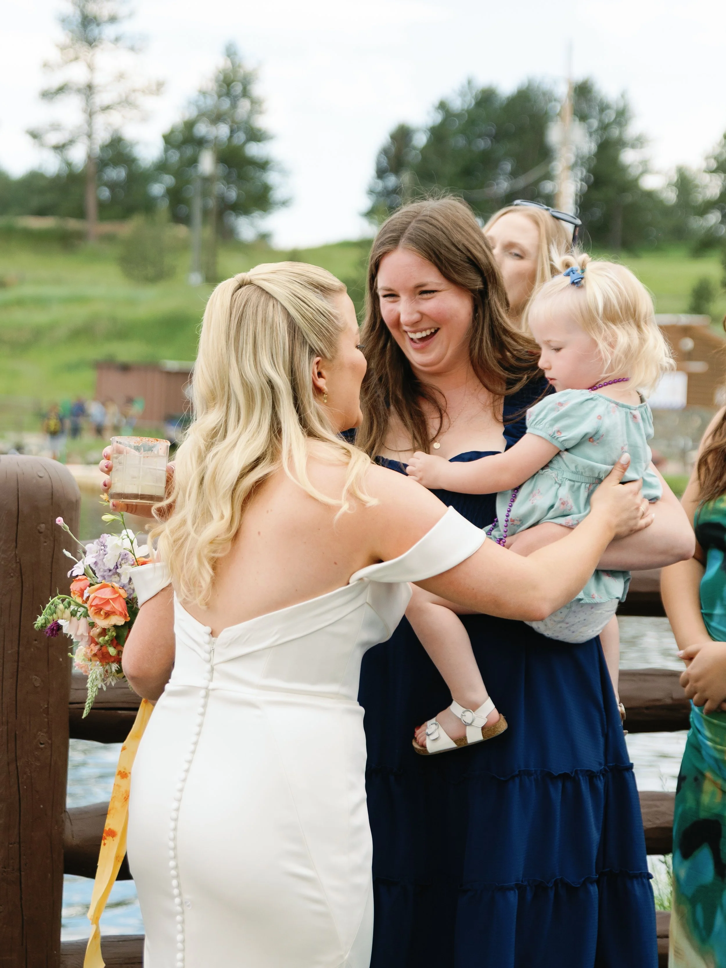 Women at a wedding embrace and smile outdoors near a lake with trees in the background. One woman wears a white dress with off-shoulder sleeves, holding a bouquet, while another woman holds a young girl wearing a mint green dress.