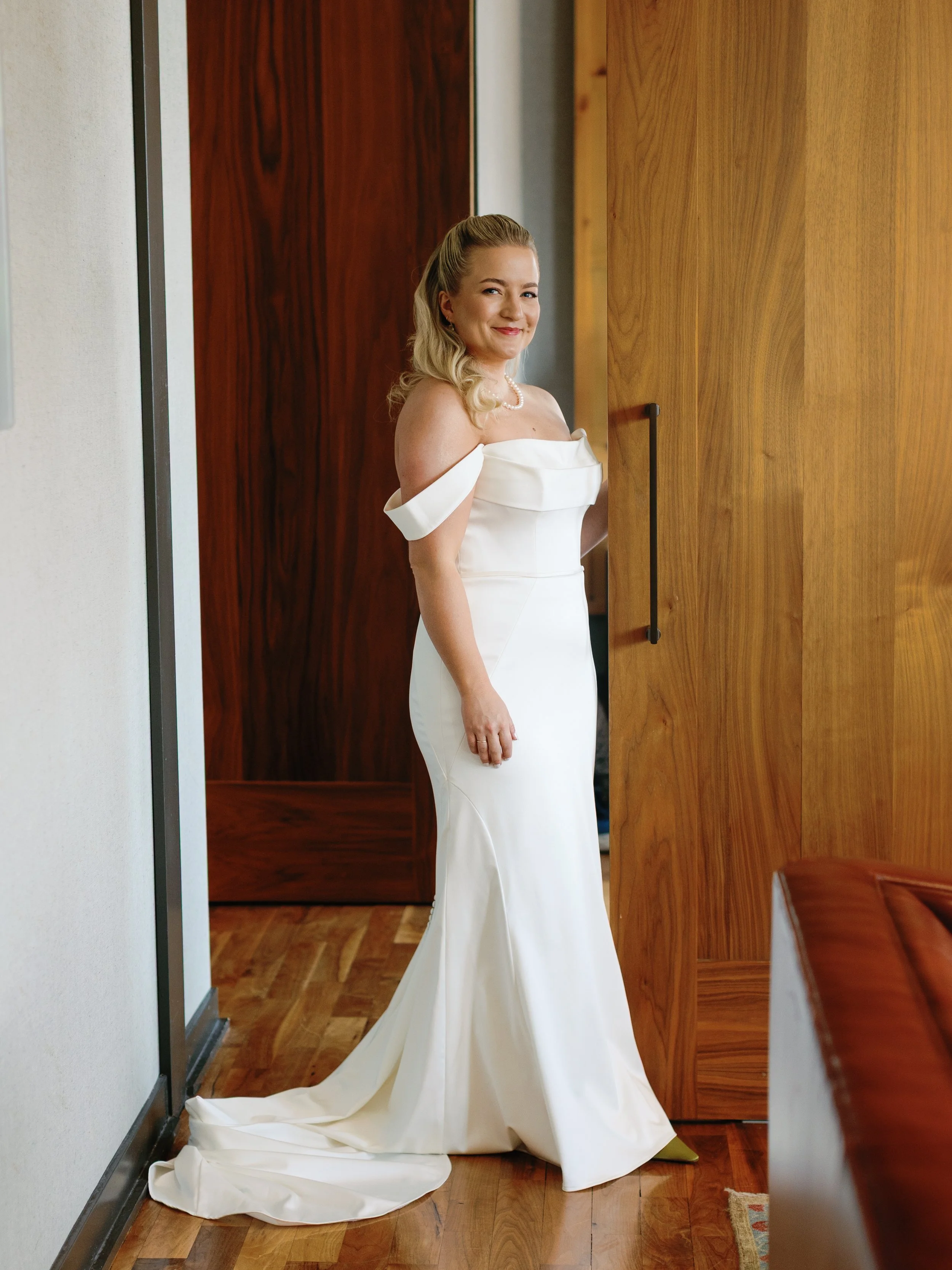 A woman in an elegant white off-shoulder wedding gown stands indoors, smiling at the camera.