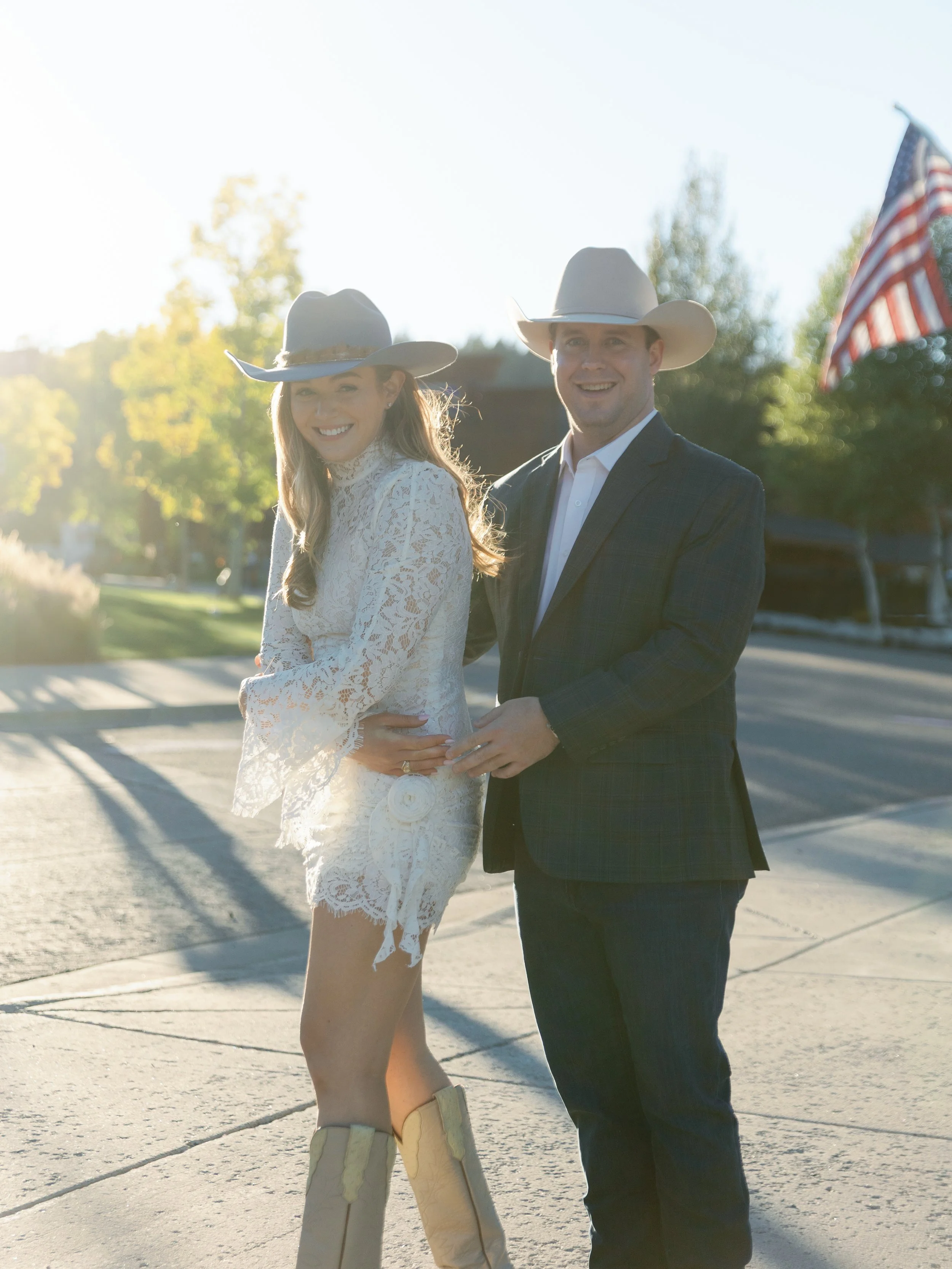 A man and woman dressed in western attire, wearing cowboy hats, standing outdoors on a sunny day with trees and an American flag in the background.