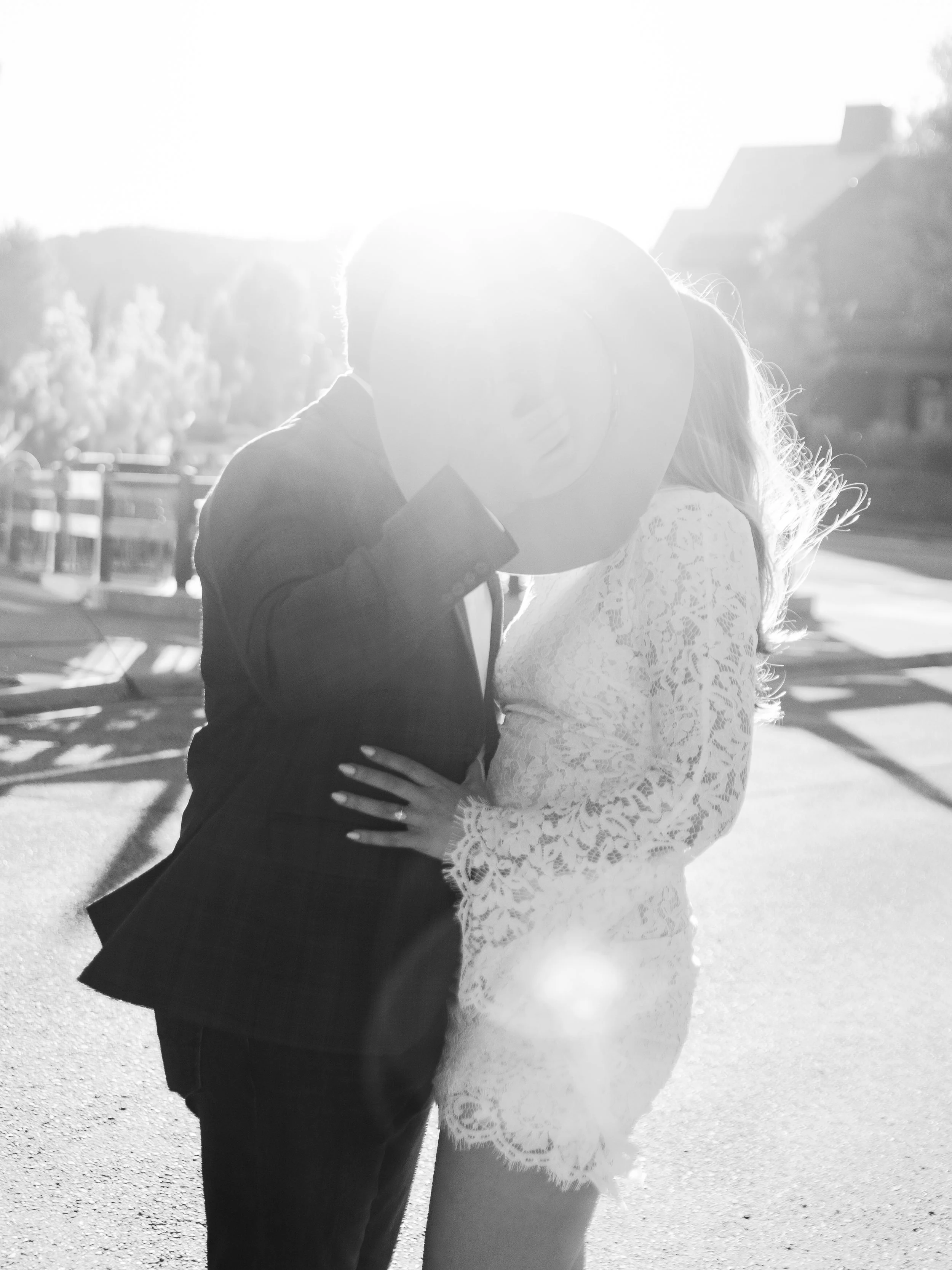 A couple standing close together outdoors, with the sun shining brightly behind them, obscuring their faces. The man is wearing a dark jacket, and the woman is in a white lace dress.
