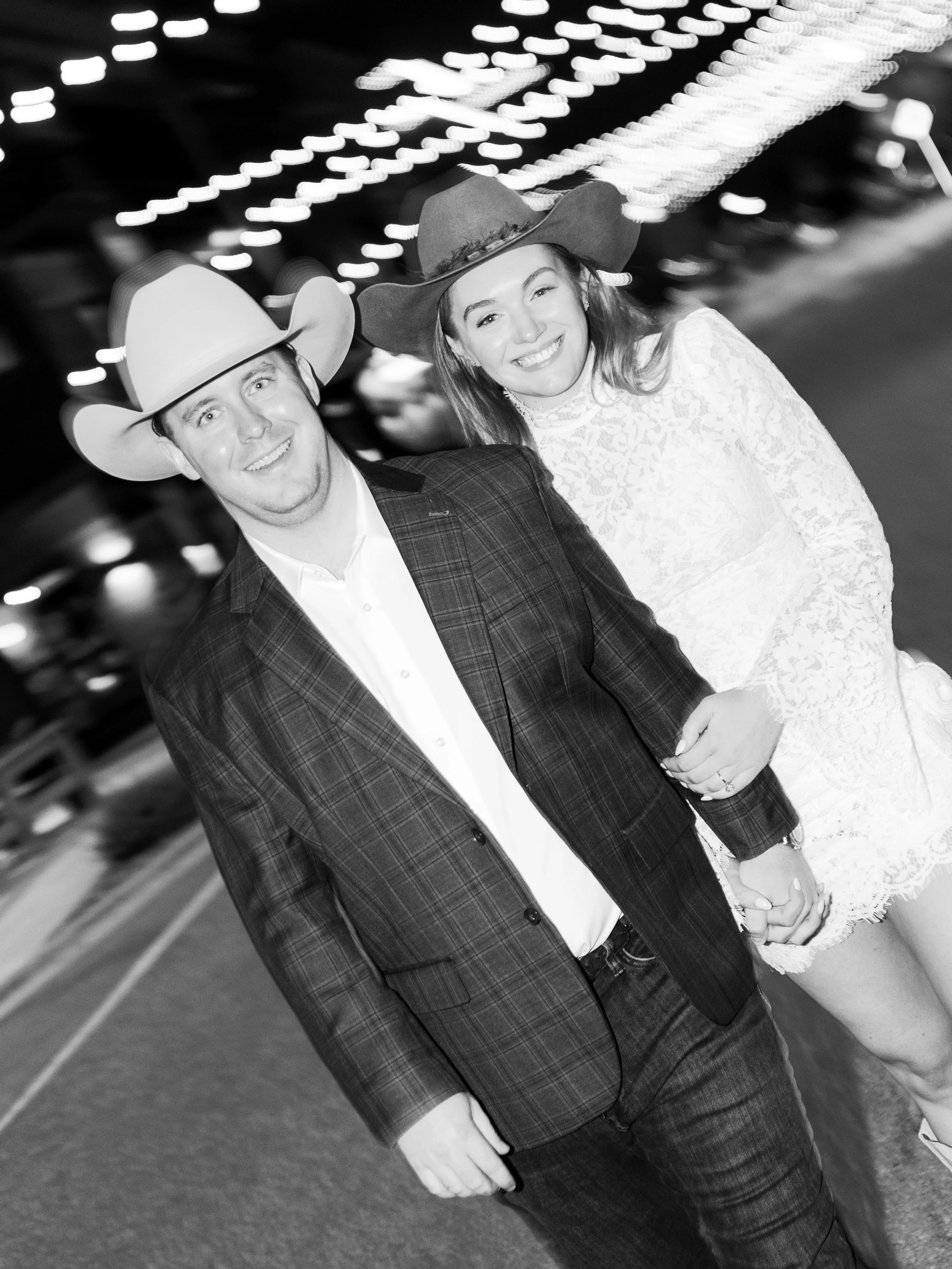 A smiling man and woman wearing cowboy hats, holding hands in a parking lot at night. The woman is in a white lace dress, and the man is in a plaid blazer with a white shirt.