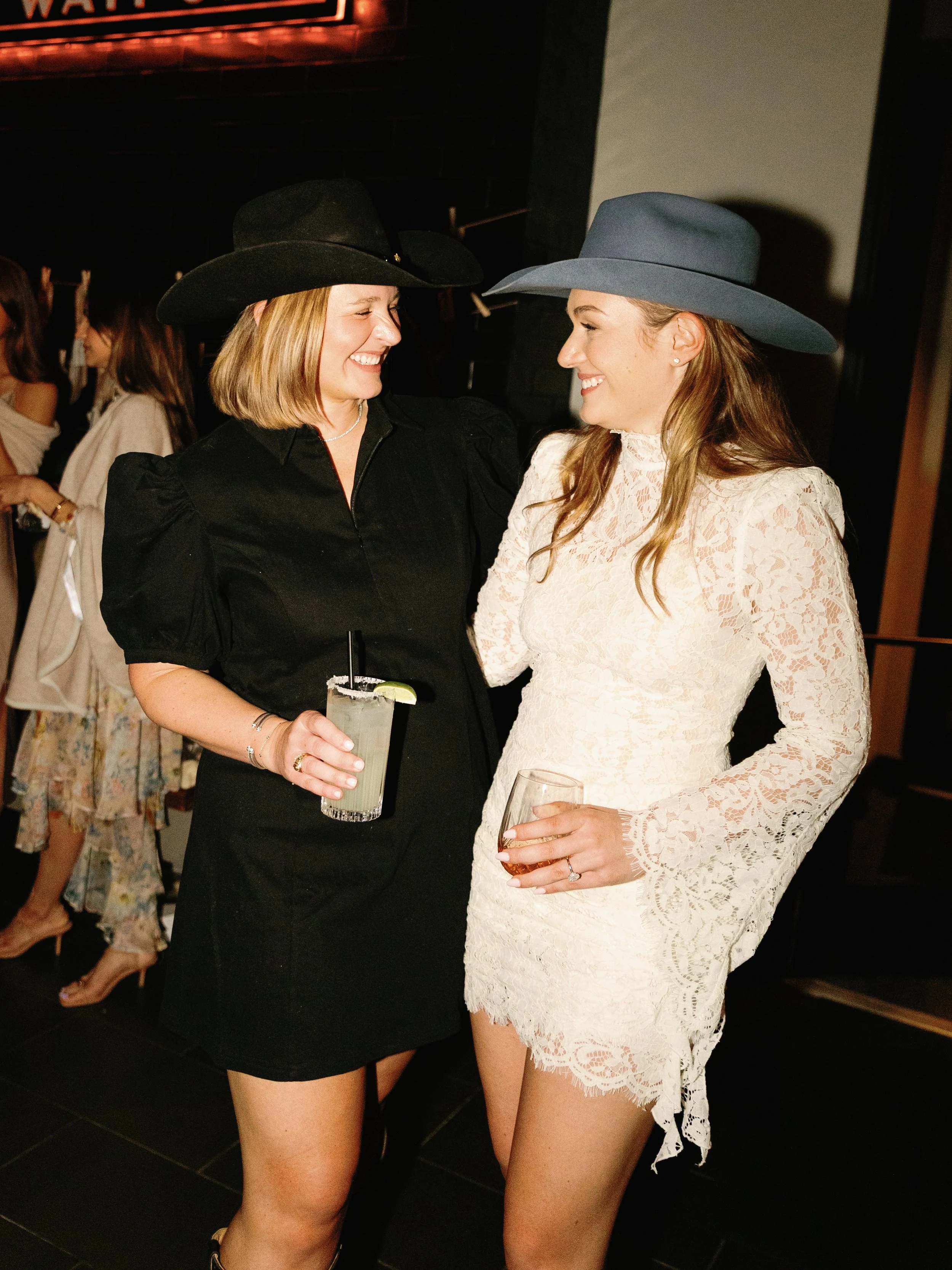 Two women in cowboy hats and dresses talking and smiling at a social event.