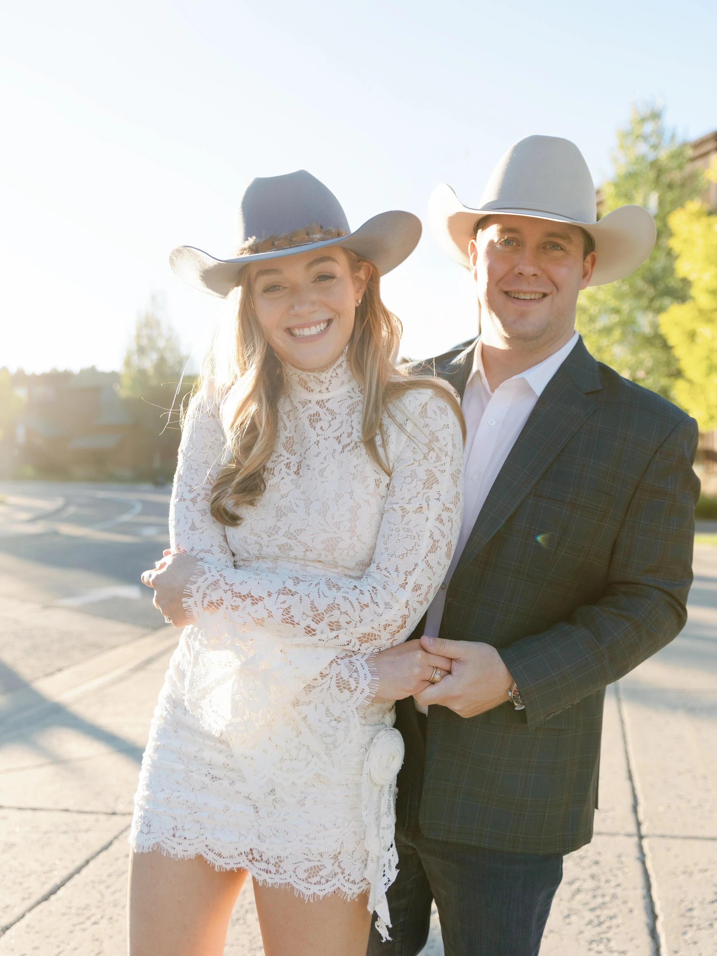 A smiling young woman and man wearing cowboy hats stand together outdoors in bright sunlight. The woman is dressed in a white lace dress, and the man is dressed in a dark plaid suit. They are holding hands and standing on a sidewalk with trees and ho