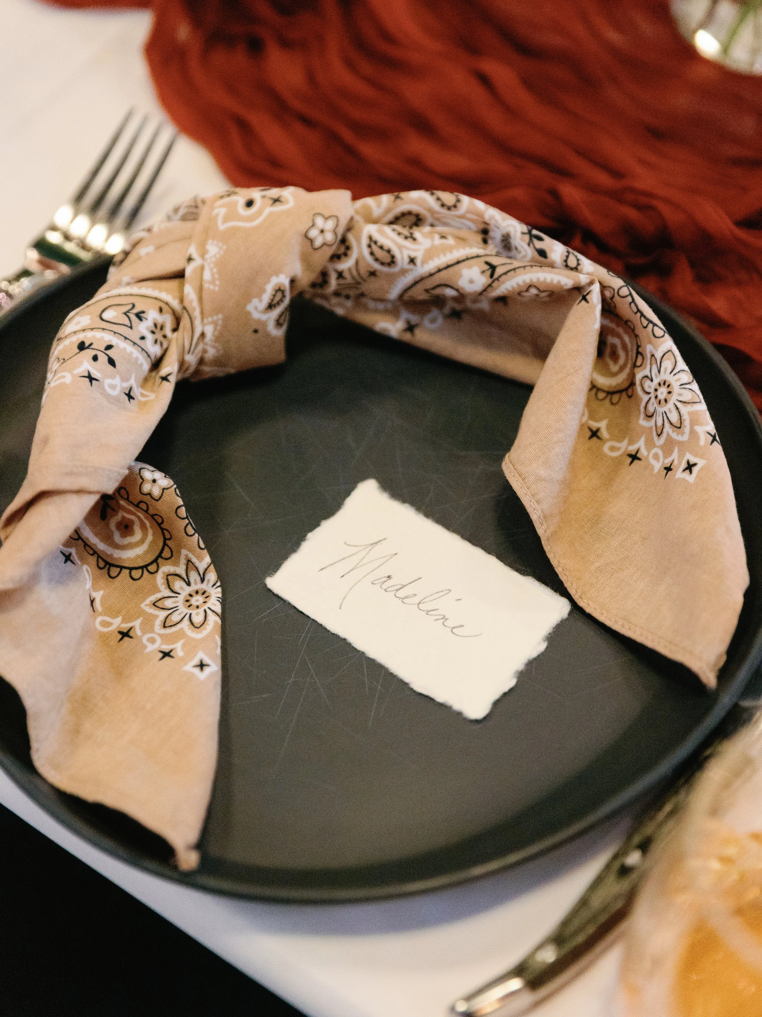 A beige bandana with a white floral pattern tied around a black plate. A torn piece of paper with the name 'Madeline' handwritten on it is placed on the plate. In the background, part of a fork and a red textured fabric are visible.