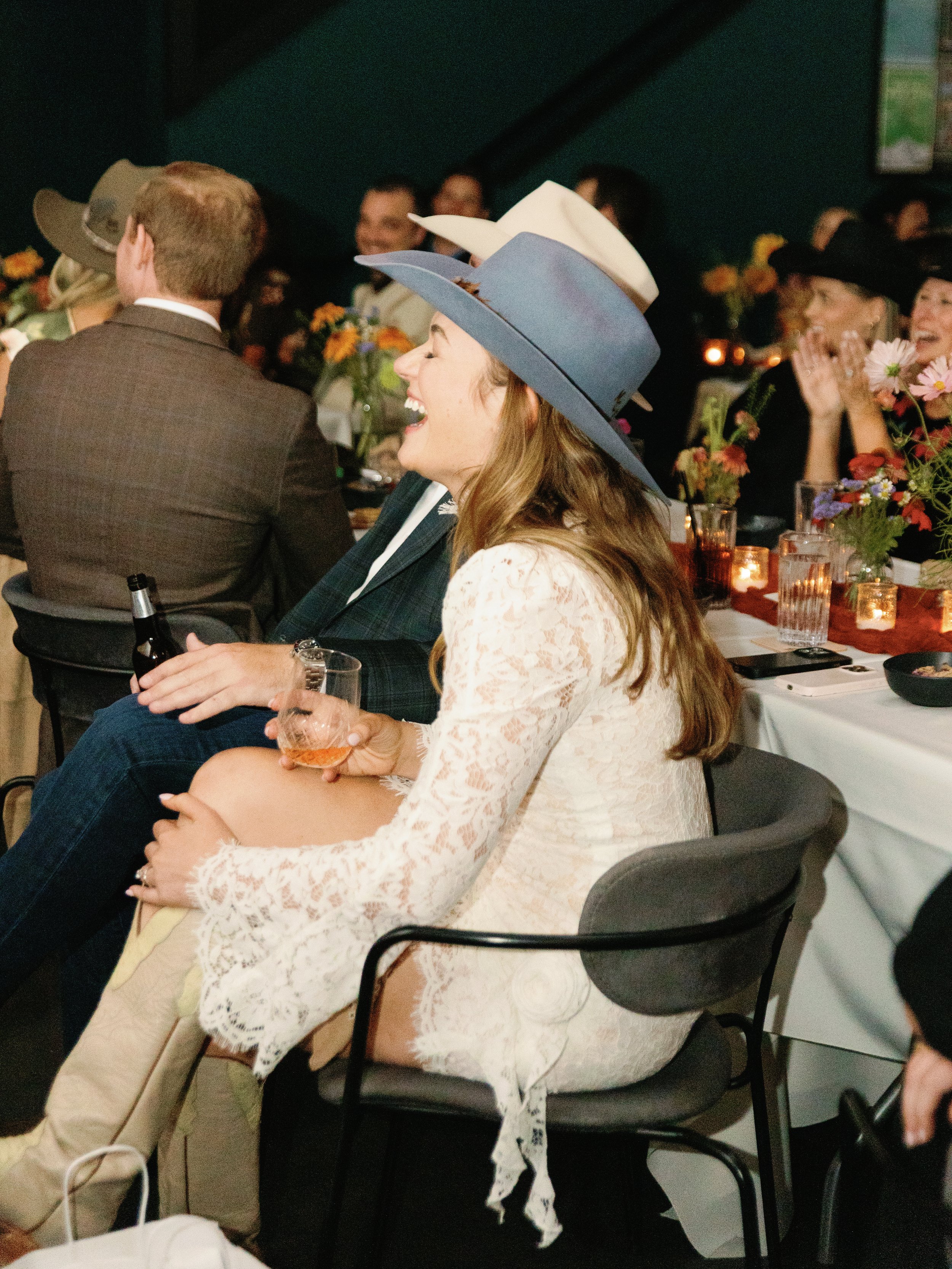 A woman with long hair, wearing a white lace dress and a large blue hat, sitting at a dinner event, holding a glass of wine, and smiling.