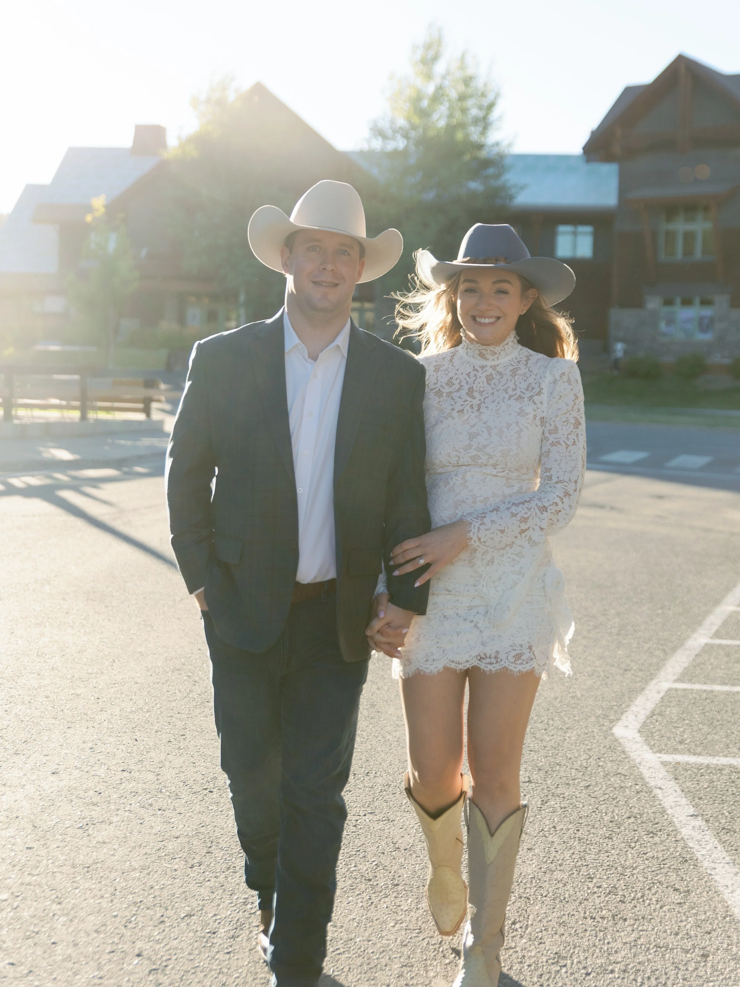 A man and woman walking hand-in-hand outdoors during sunset, both wearing cowboy hats and Western-style clothing, smiling, with houses and trees in the background.
