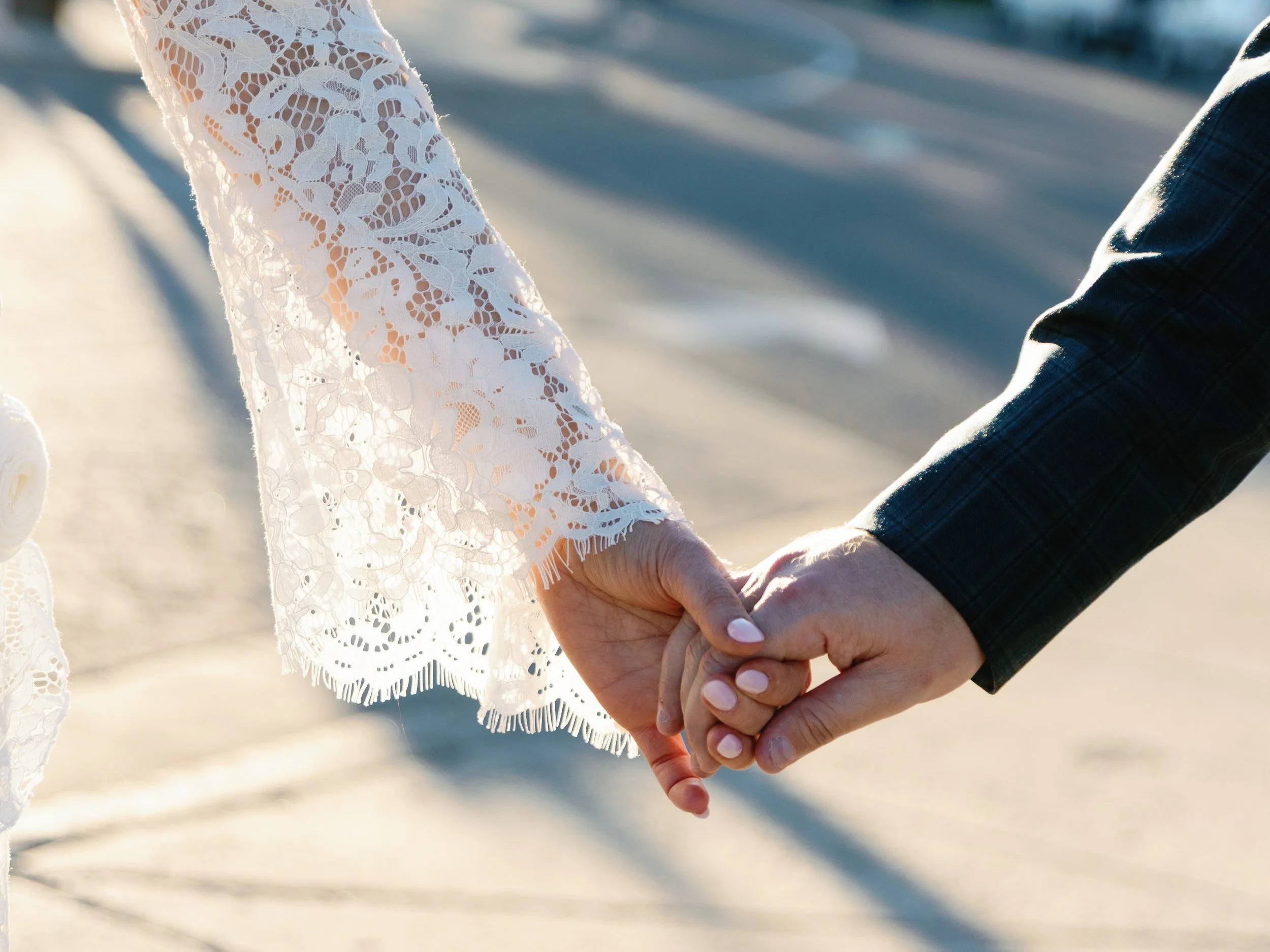 A couple holding hands on the beach, the woman wearing a white lace dress and the man in a dark suit, with sunlight casting a warm glow.