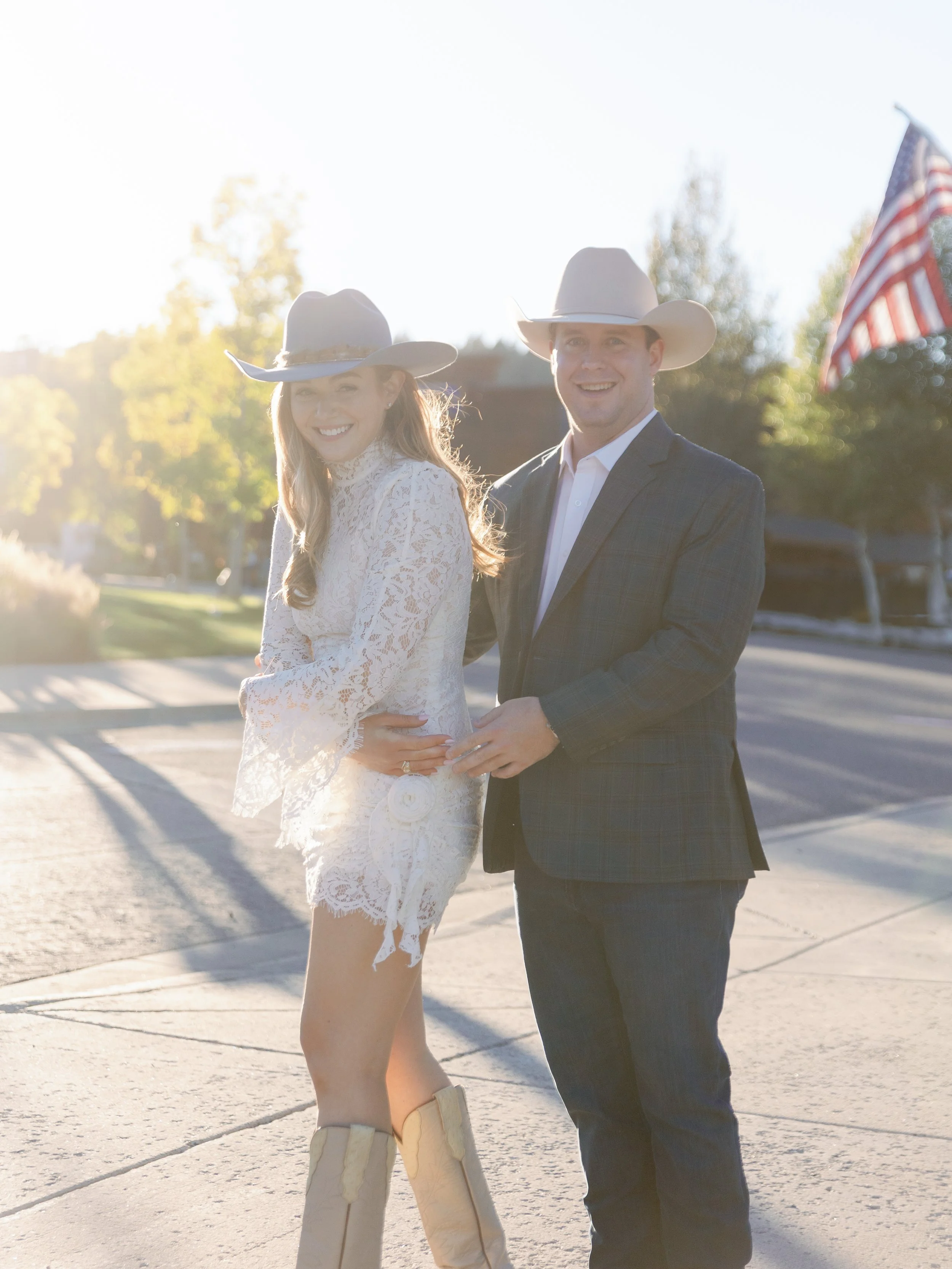 A smiling couple dressed in Western attire, wearing cowboy hats, standing outdoors during golden hour.