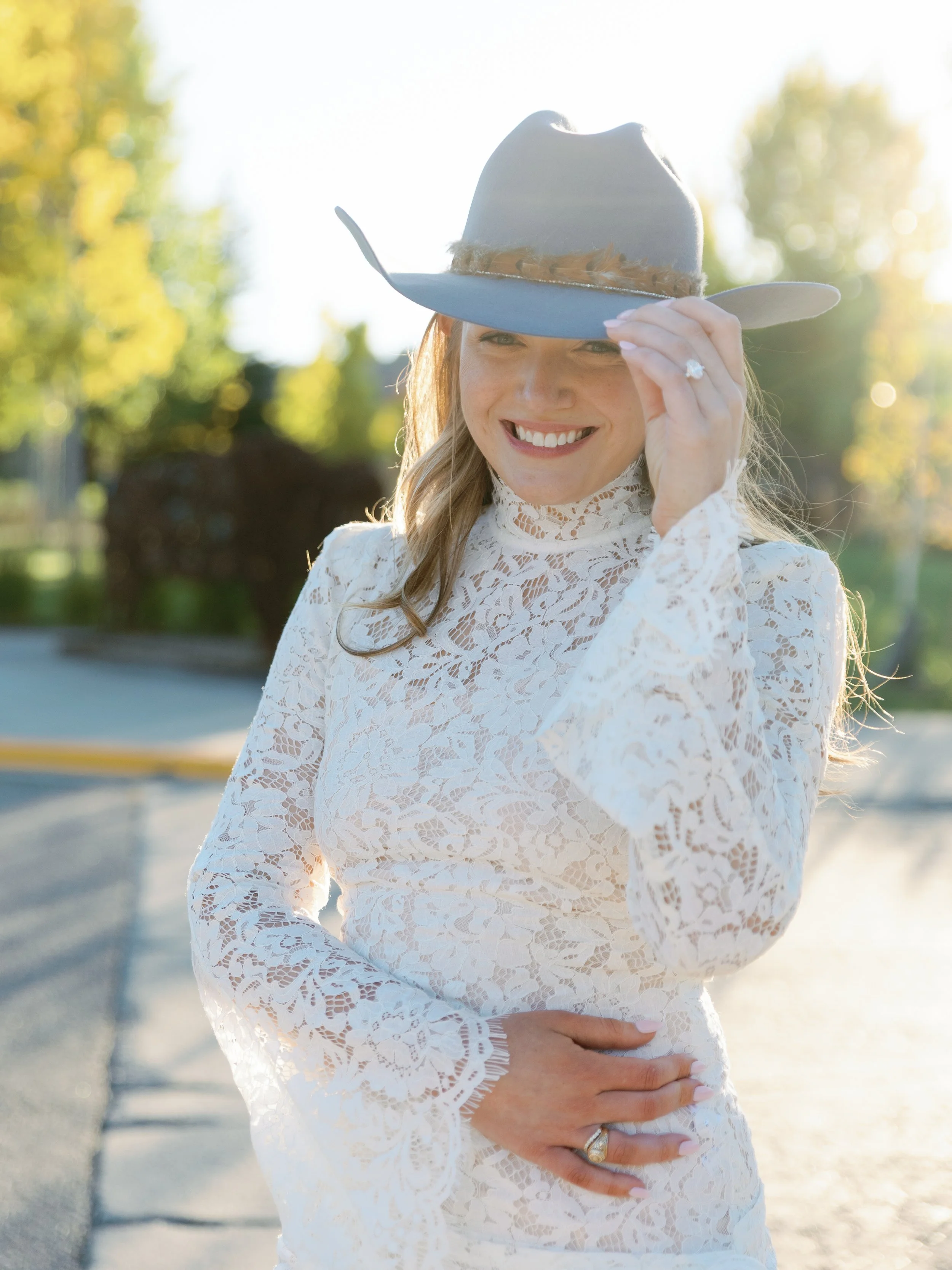 A woman in a white lace dress and a gray cowboy hat with a feather, smiling outdoors in sunlight.