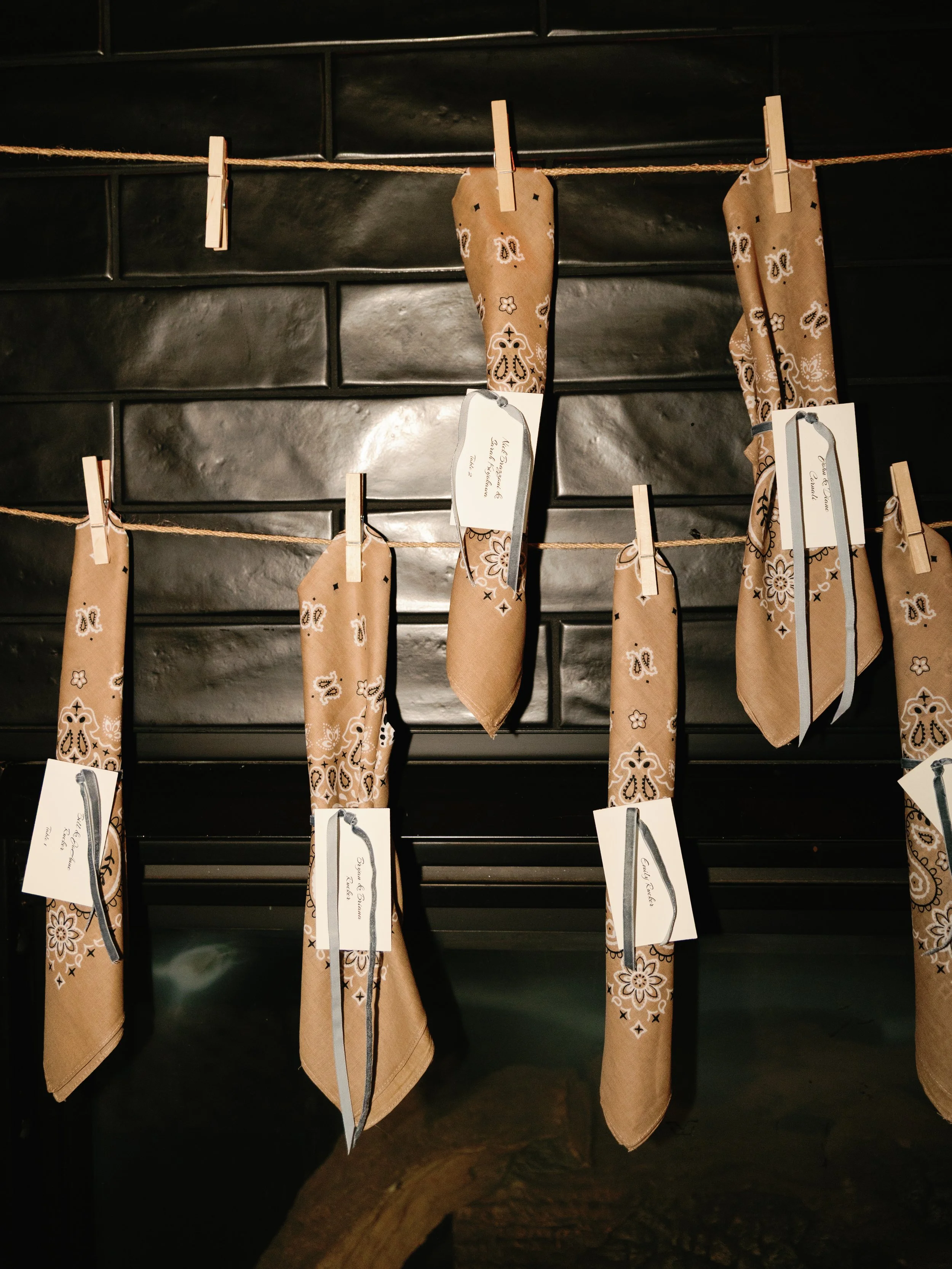 Multiple brown bandanas hanging on a string with clothespins, each with a tag attached, against a black tiled wall.