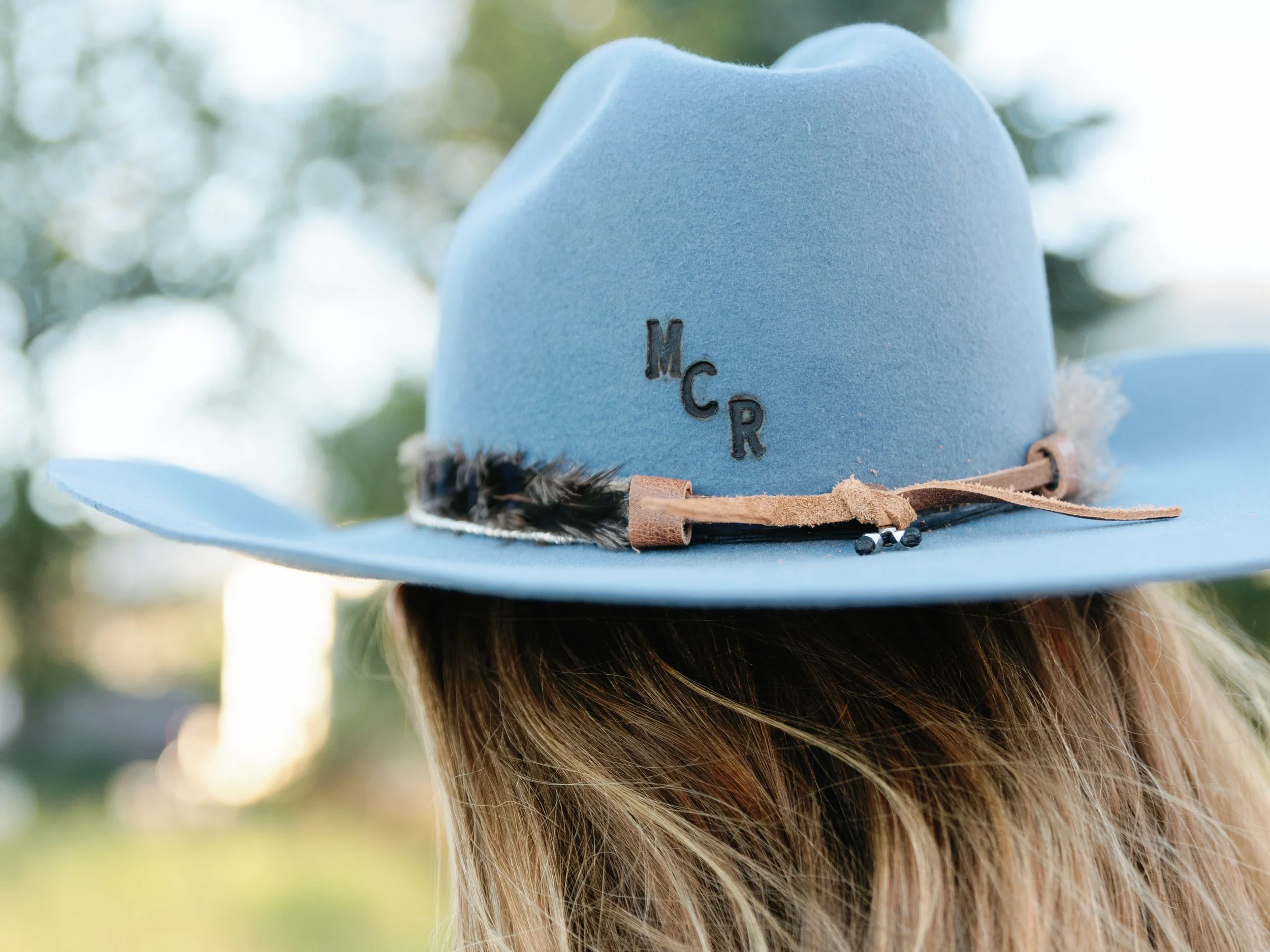 A person wearing a light blue felt wide-brimmed hat with the initials 'MCR' embroidered on the front, decorated with a leather band, feathers, and small beads, outdoors with blurred green foliage in the background.