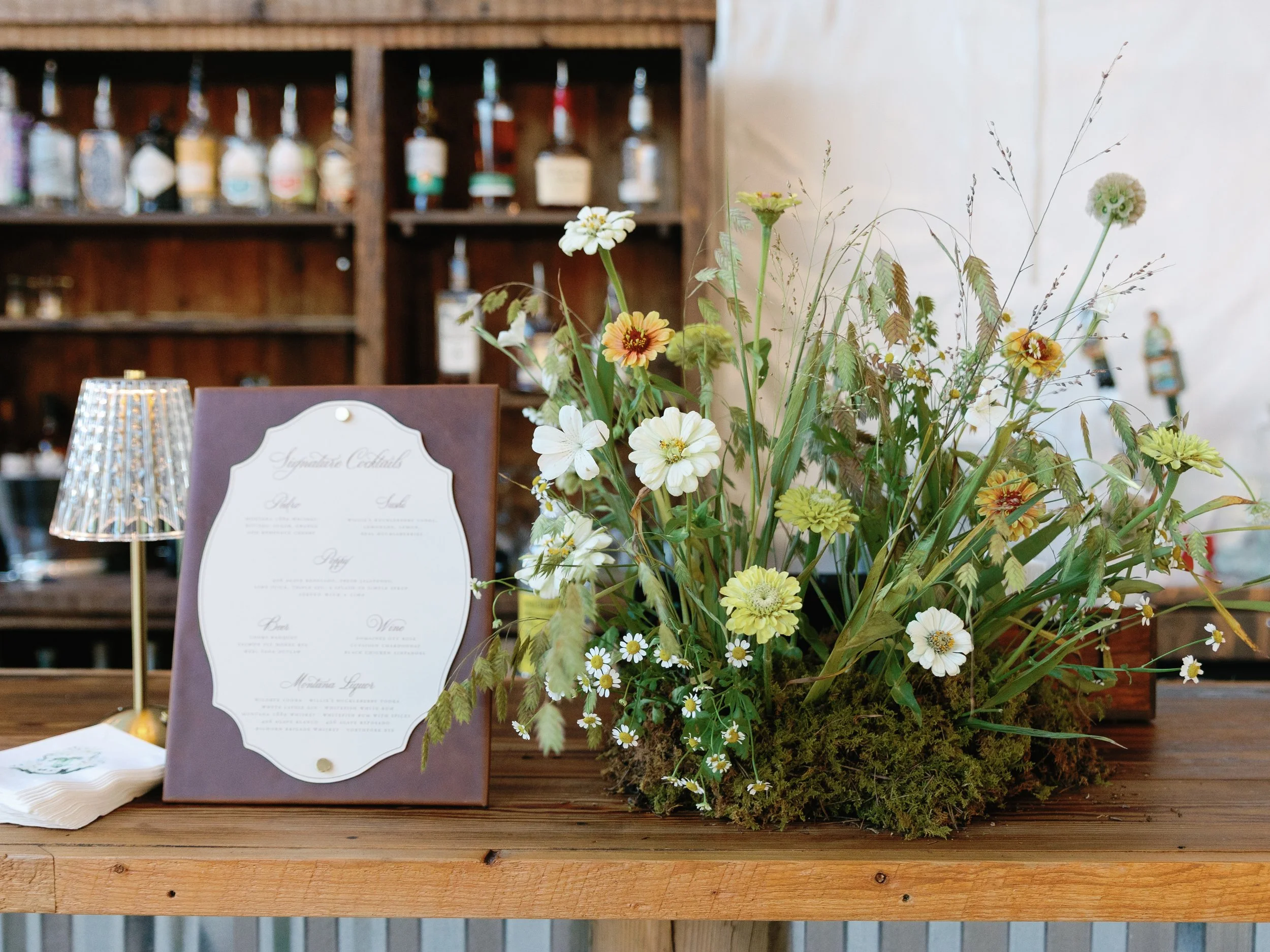 A table with a floral arrangement of white, yellow, and orange flowers and greenery, plus a menu in a folded stand and a small lampshade, with a wooden bar and liquor bottles in the background.