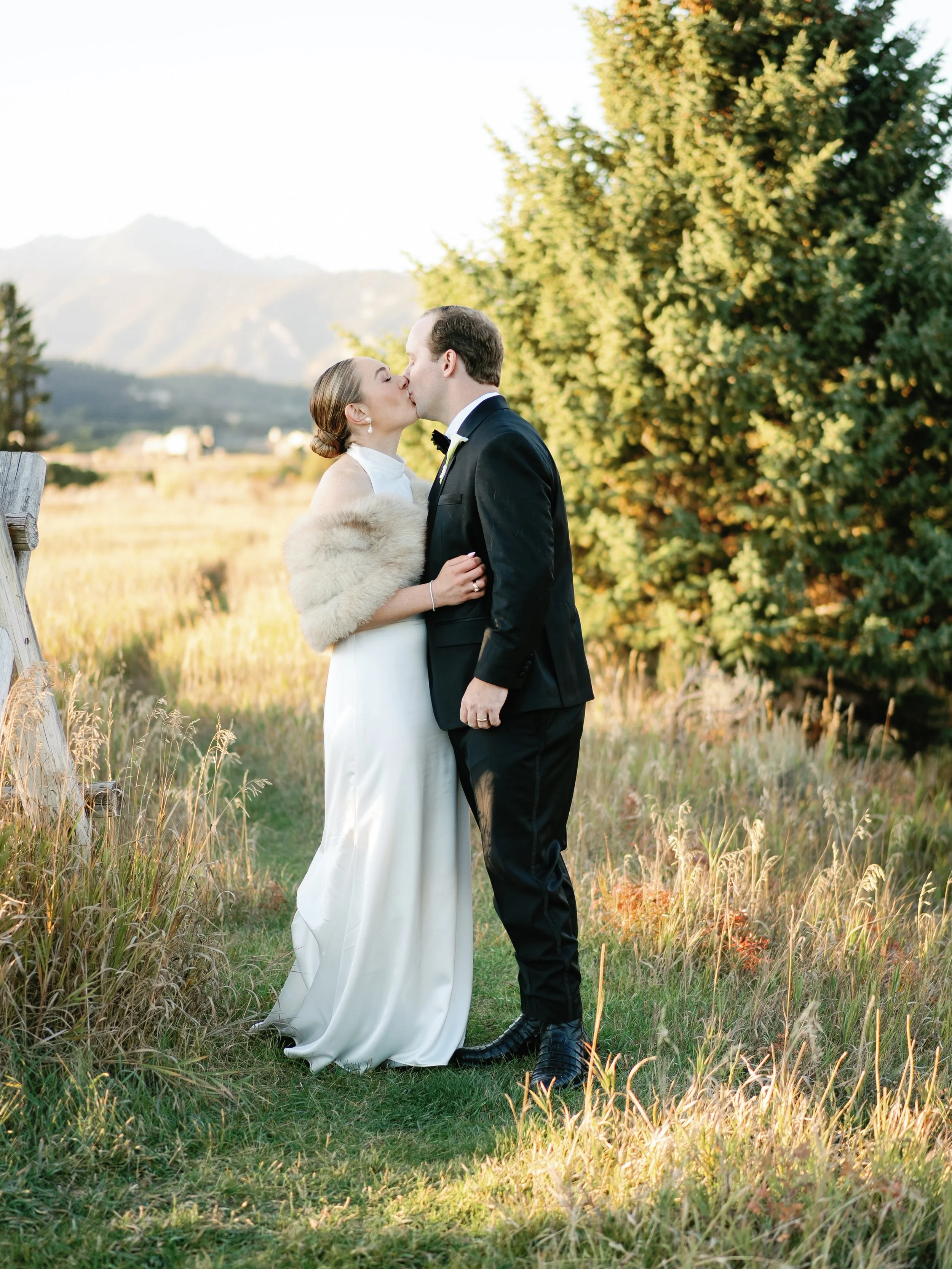A bride and groom kiss outdoors in a grassy field with mountains and trees in the background, during what appears to be a wedding.
