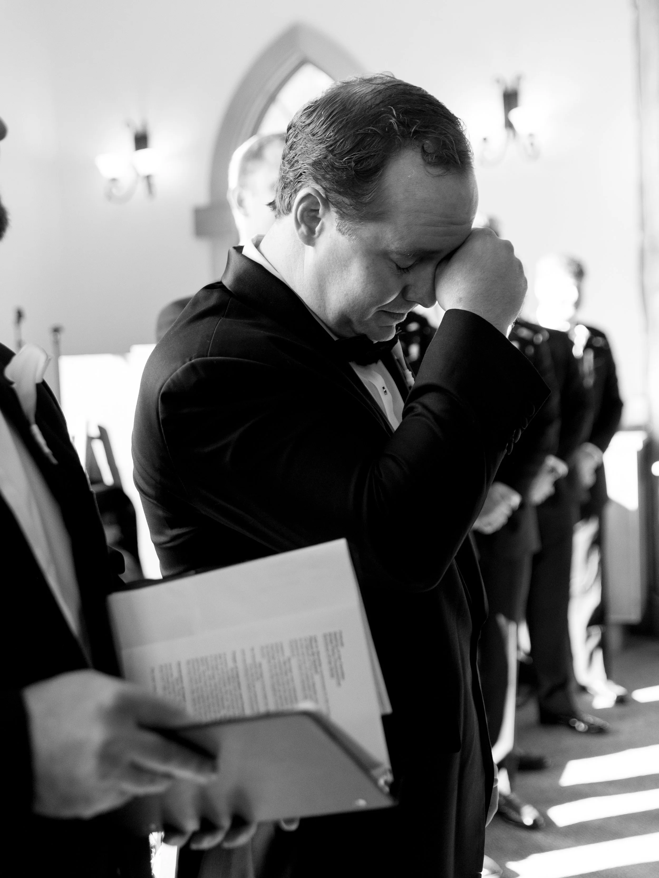 A man in a tuxedo and bow tie prays with his eyes closed and hand to his forehead during a formal ceremony, with people standing in line behind him.