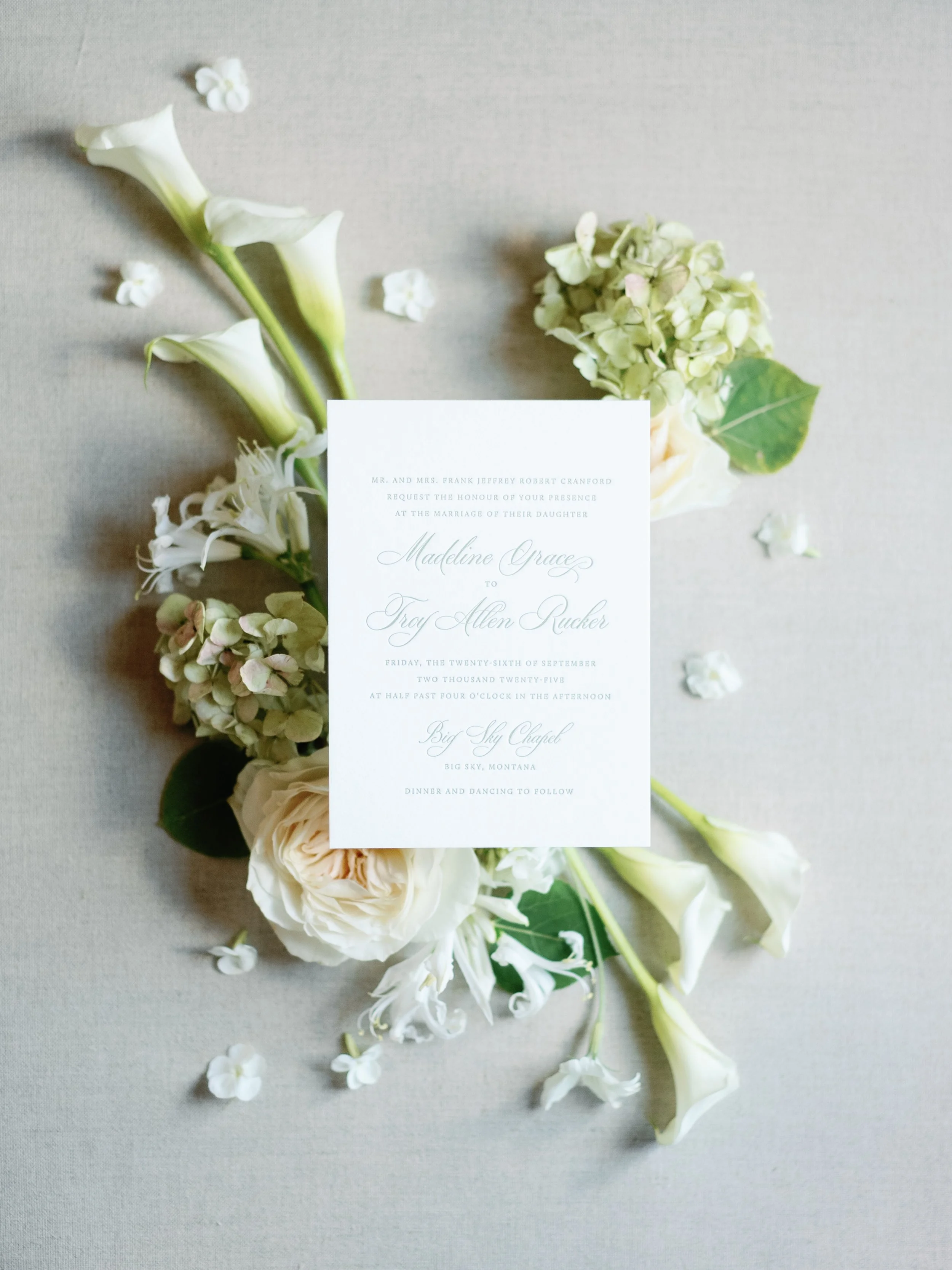 Wedding invitation card decorated with white flowers and green leaves, lying on a light-colored surface.