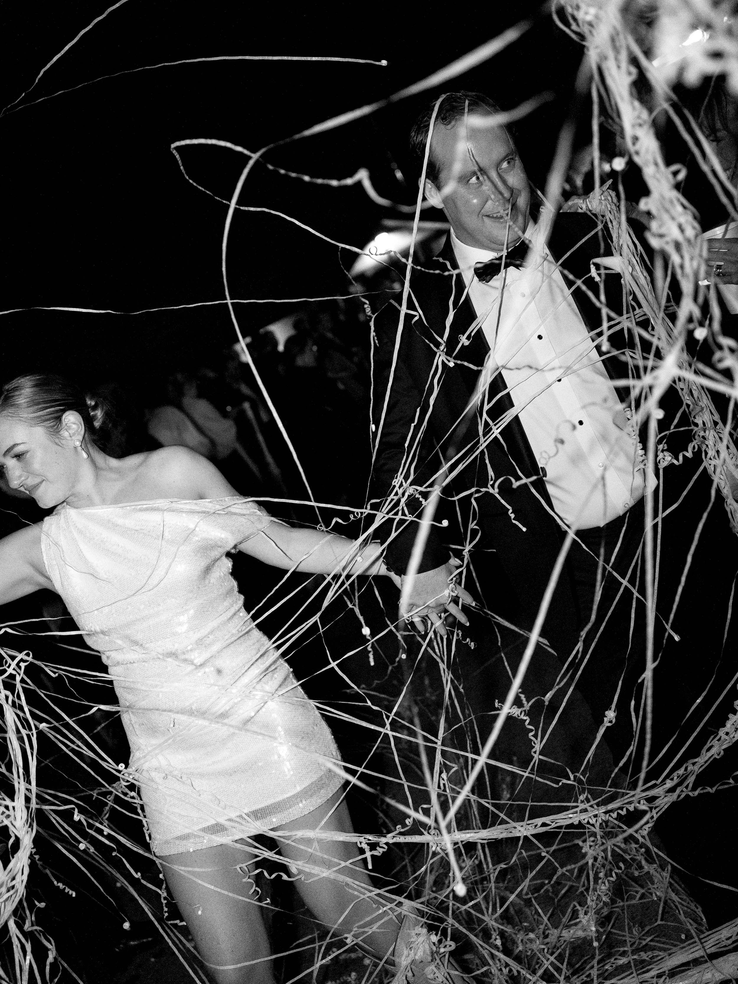 A bride and groom dancing at a wedding reception, holding hands, surrounded by streamers and decorations.