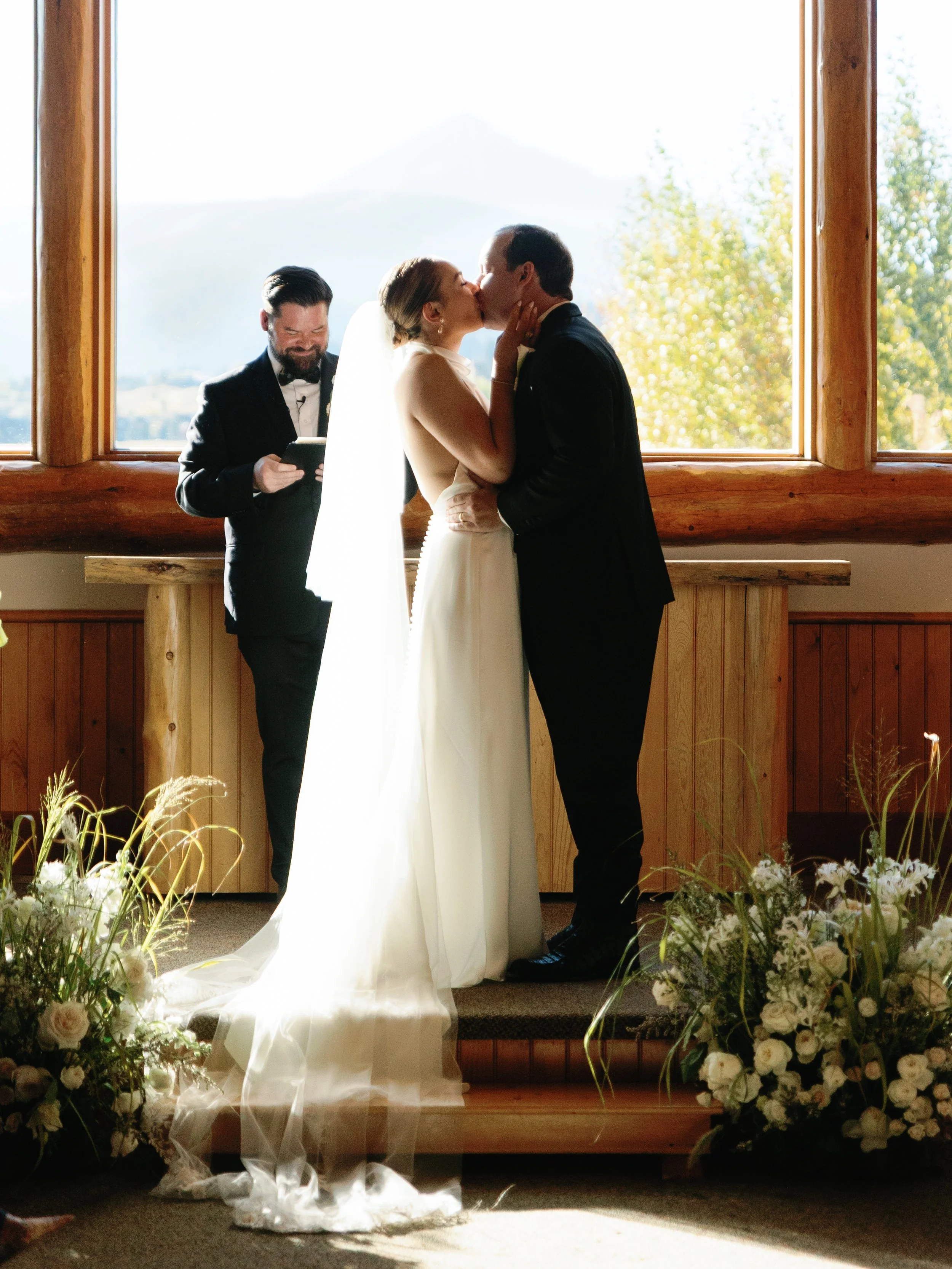 A bride and groom kiss during their wedding ceremony in front of a large window with a mountain view. An officiant stands behind them, holding a book.