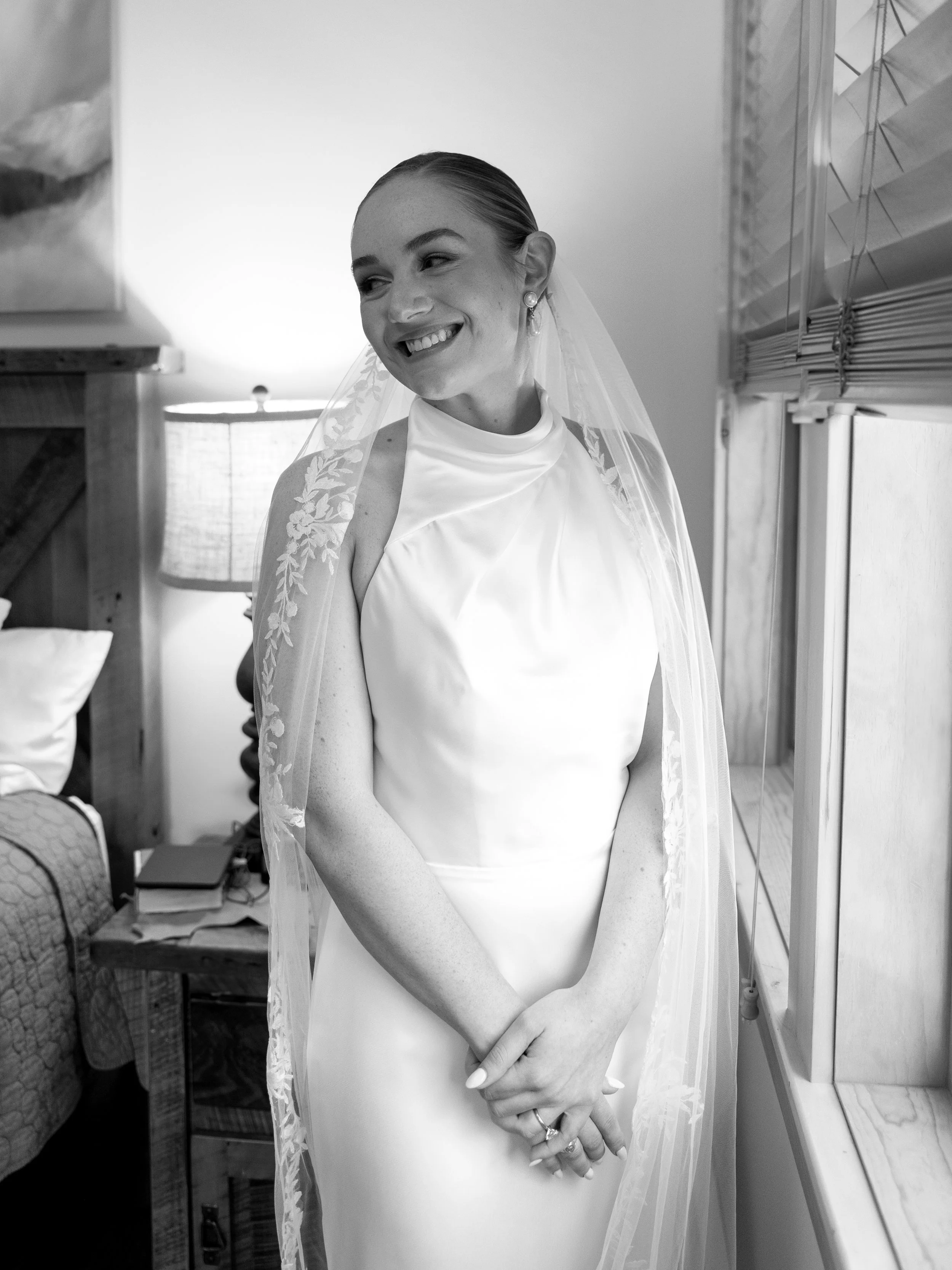 A bride stands indoors in a room with wooden furniture, wearing a satin wedding dress with a high neckline and lace veil, smiling while looking to her left.