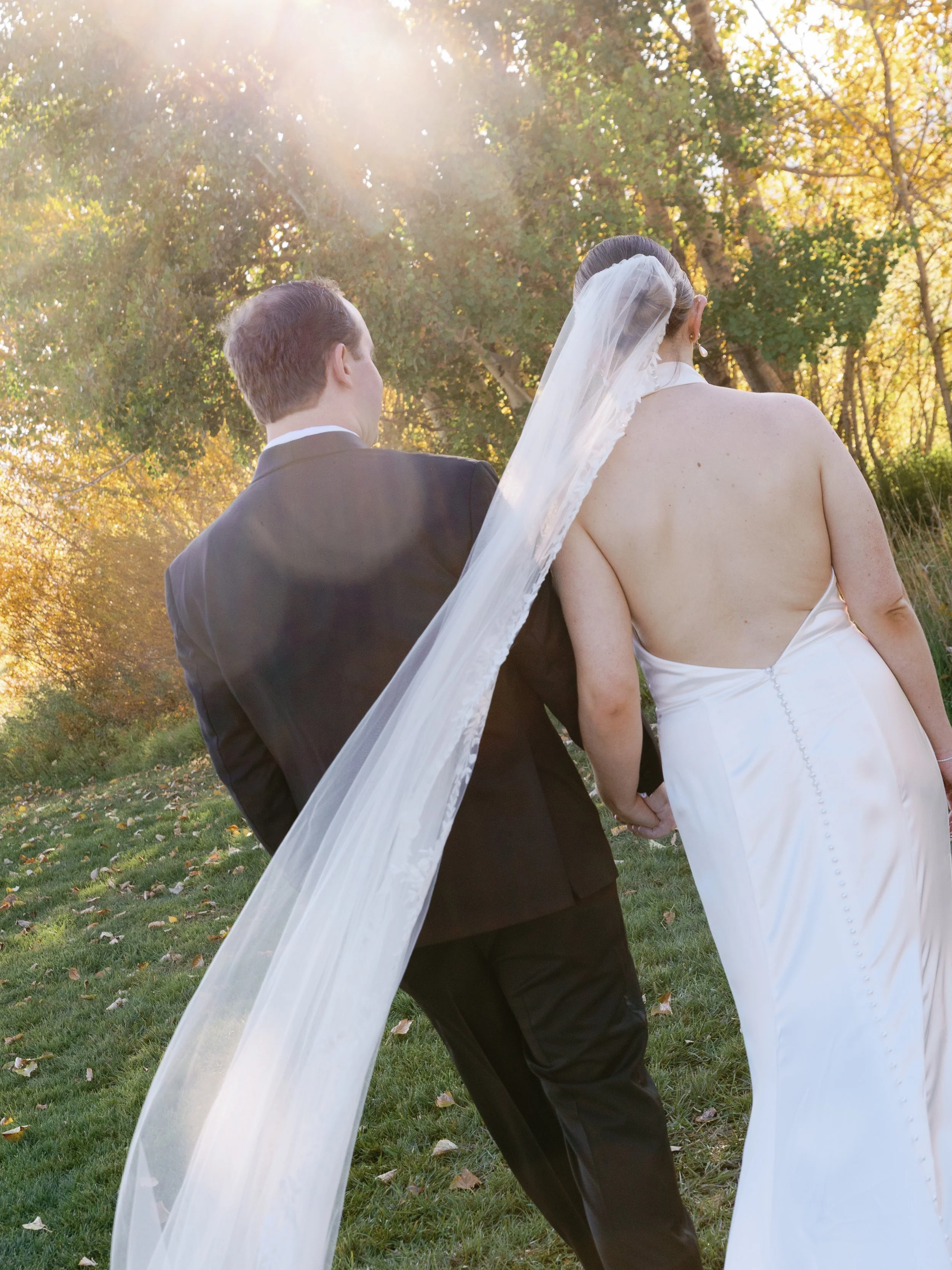 A bride and groom holding hands outdoors, walking away from camera with sunlight shining through trees in the background.
