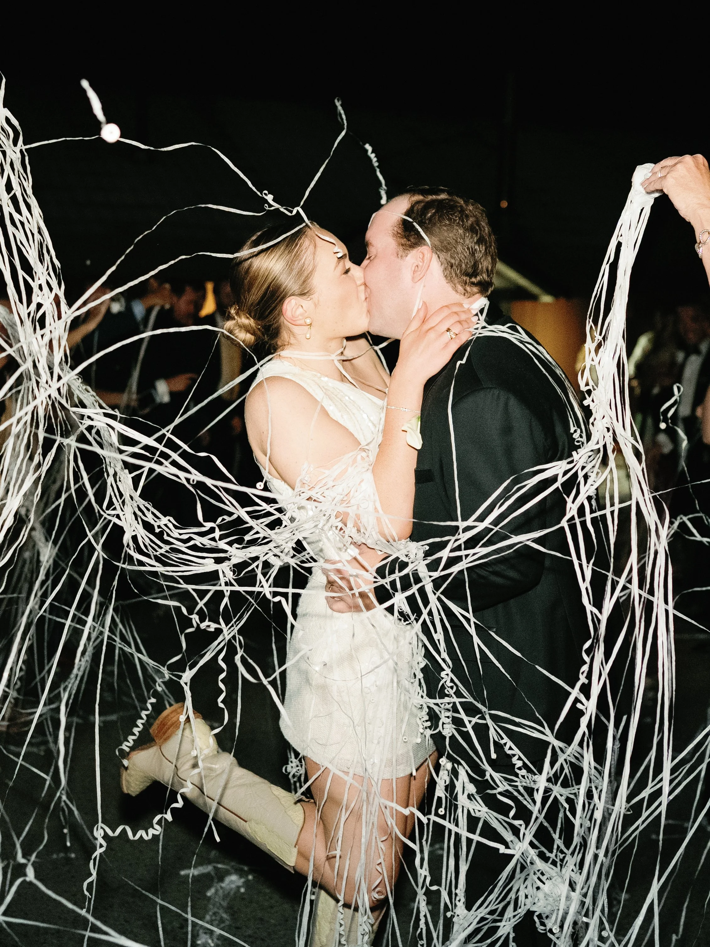 A newlywed couple kissing amid a shower of white streamers at their wedding reception.