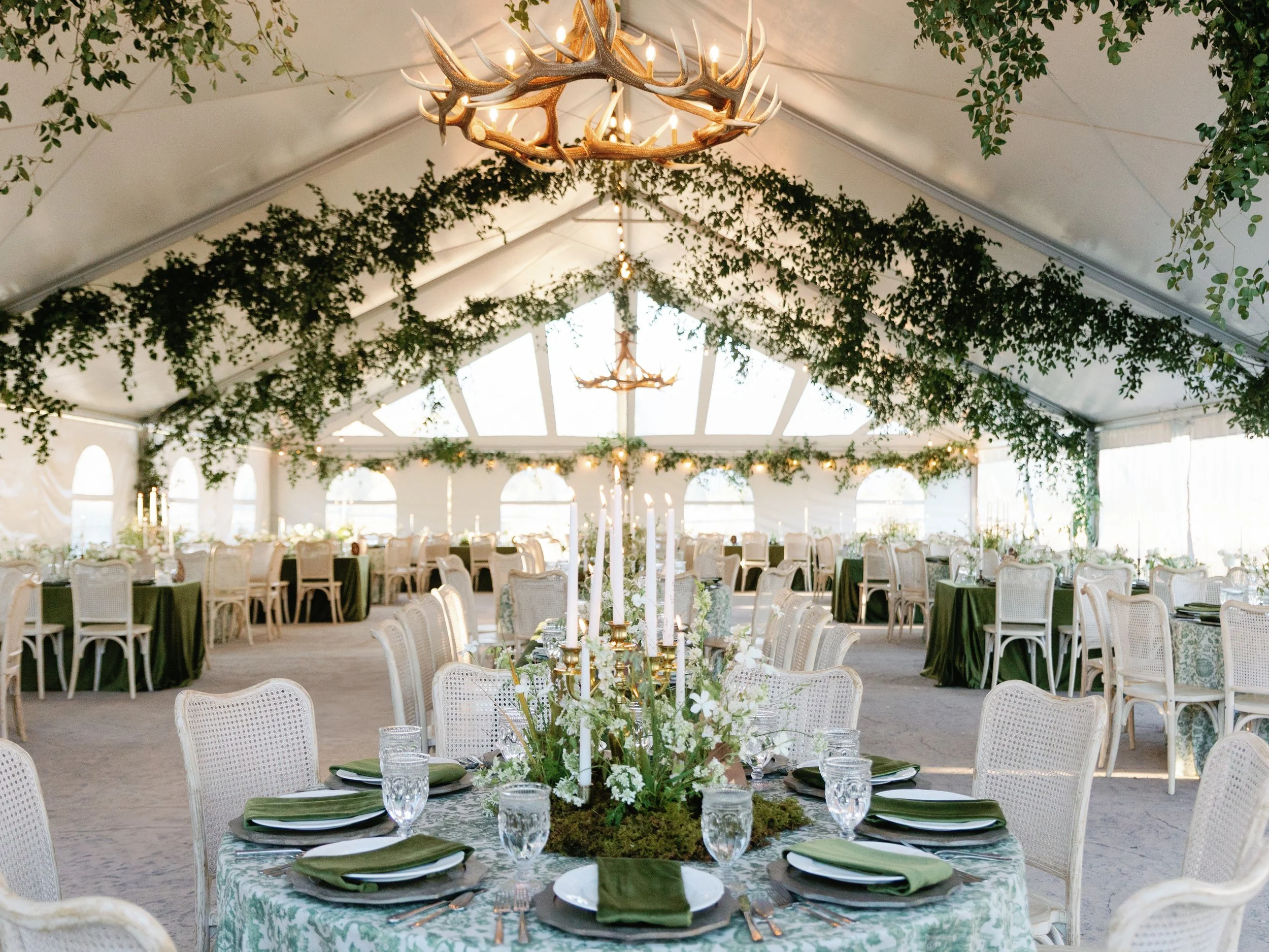 Elegant indoor wedding or event reception tent with green and white decor, floral centerpieces, candle arrangements, and chandelier-style lighting, with multiple round tables and chairs arranged for guests.