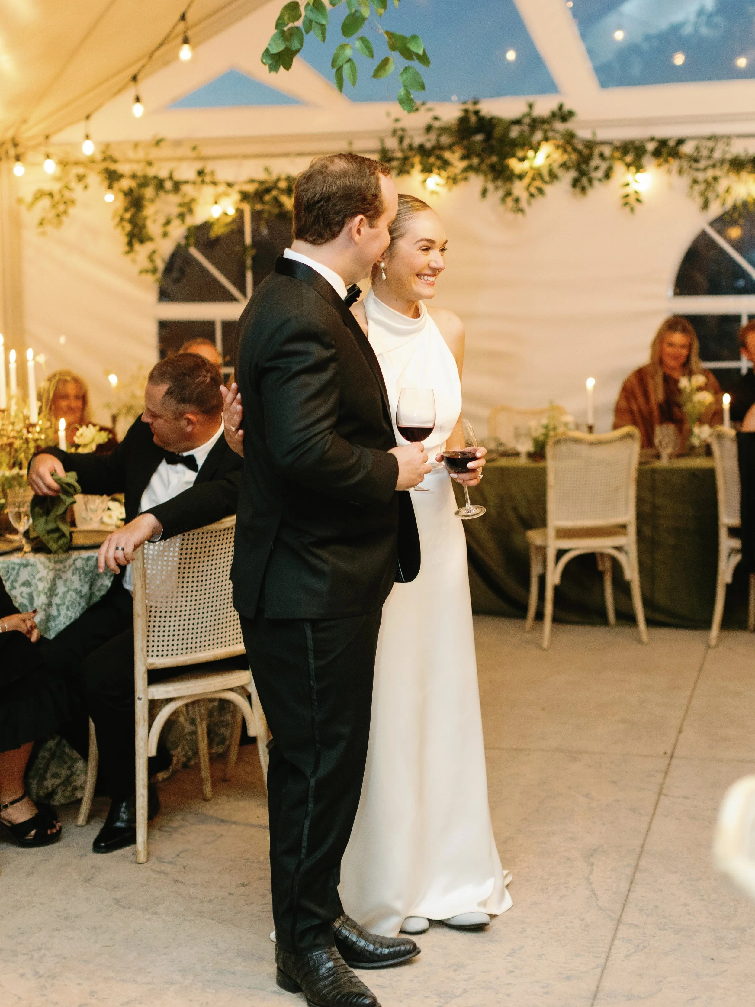 A couple at a wedding reception, dressed in formal attire, smiling and holding glasses of red wine. The man is wearing a black tuxedo with a bow tie, and the woman is in a white wedding gown. The reception is inside a decorated tent with hanging stri