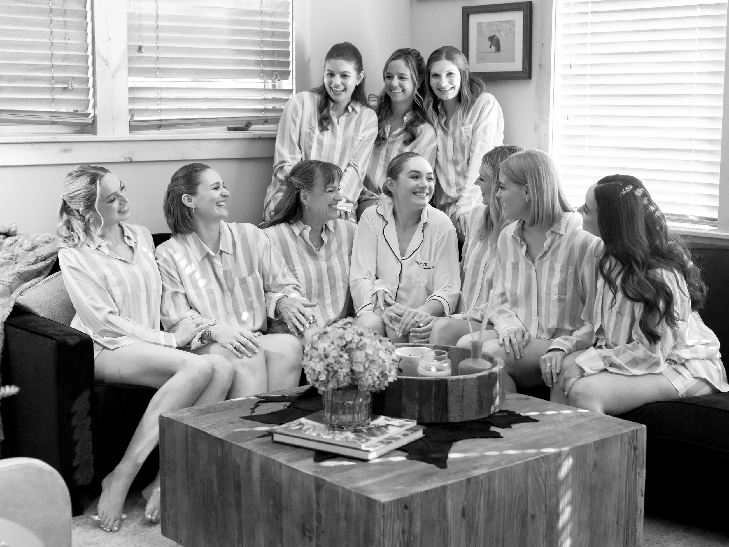 A group of women in pajamas, some sitting on a sofa and some leaning against a wall, laughing and conversing in a cozy living room, with a coffee table holding flowers and candles in front of them.