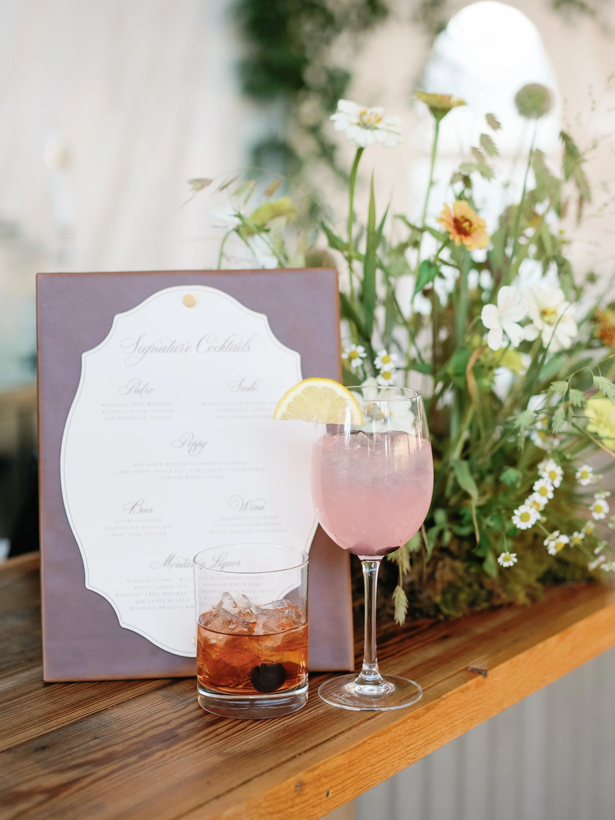 A pink cocktail with a lemon wedge on a glass rim, a dark drink with ice and a cherry in a short glass, and a floral arrangement of mixed white and yellow flowers on a wooden surface with a light-colored background.