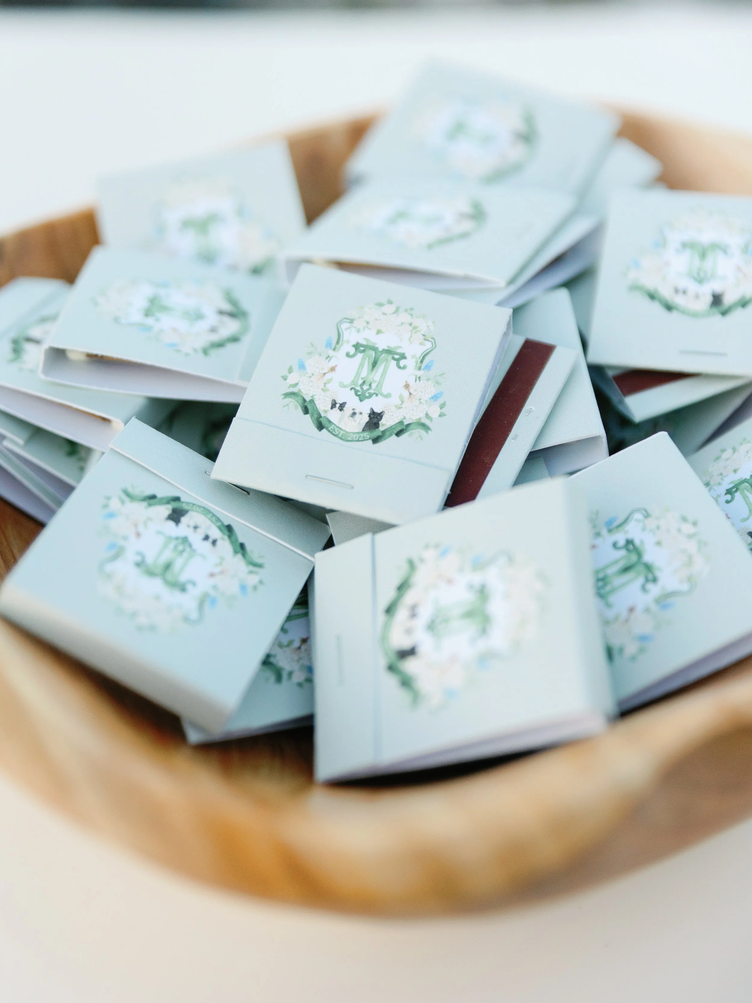 A wooden bowl filled with small light blue boxes with floral designs and a monogram, used for wedding favors or decorations.